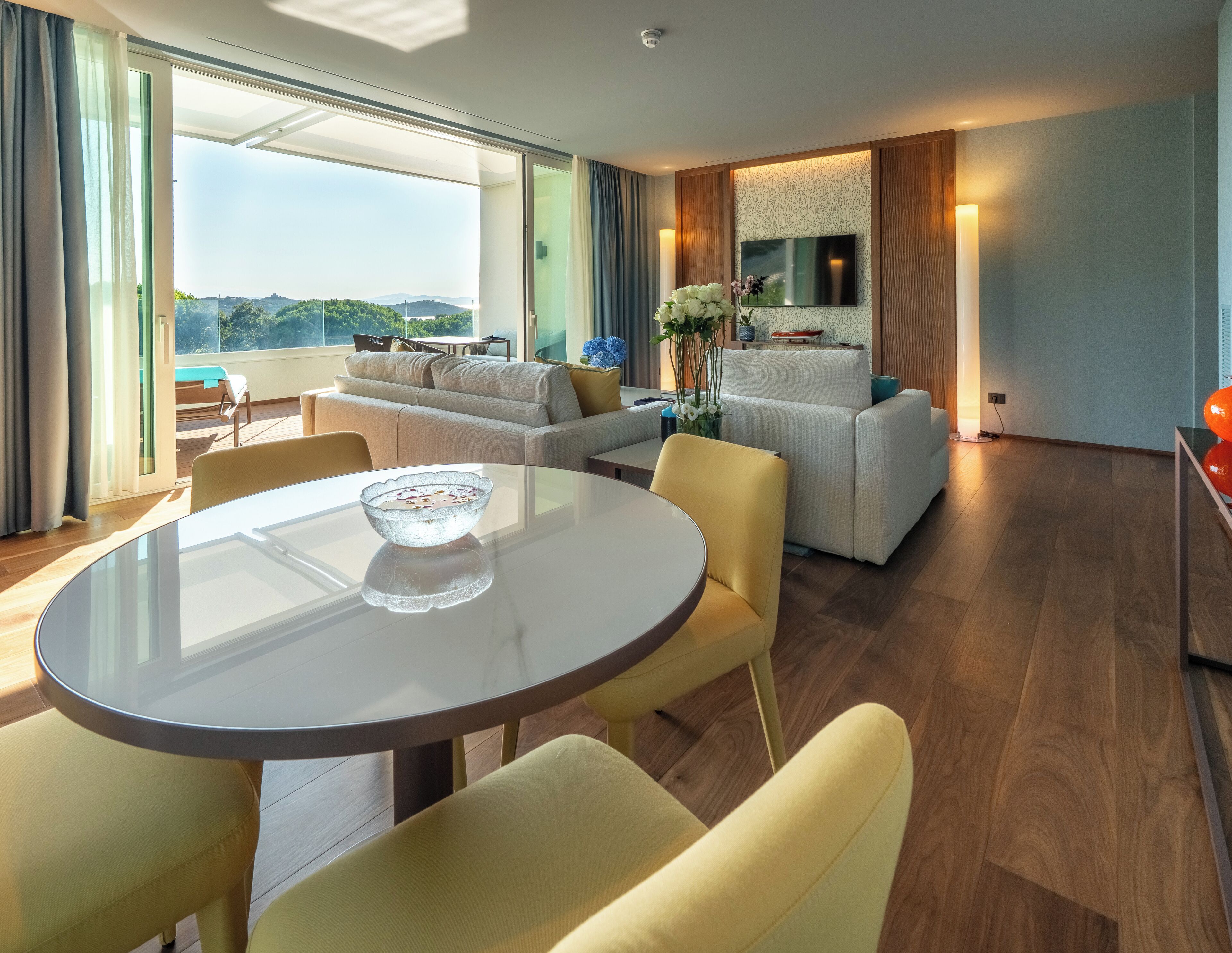 Una moderna camera d'hotel con un'ampia finestra, una zona pranzo con tavolo in vetro e un salotto con divani e televisore.