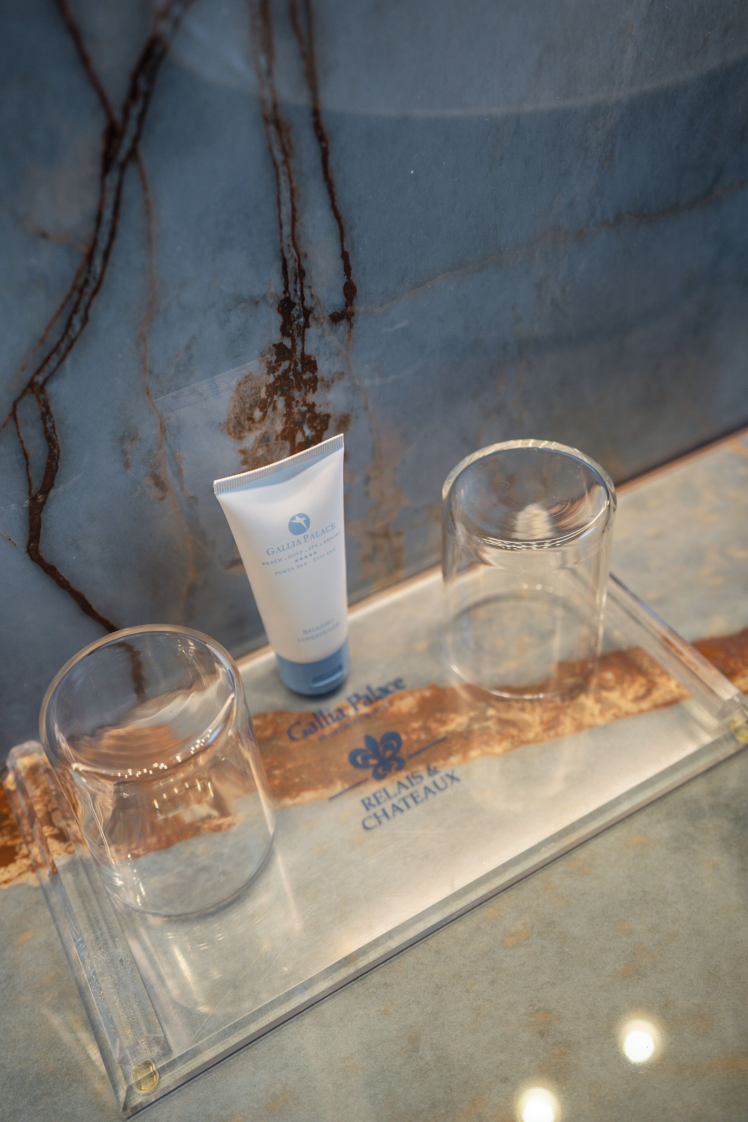 A bathroom counter with a marble wall, a tube of lotion, and two glasses.