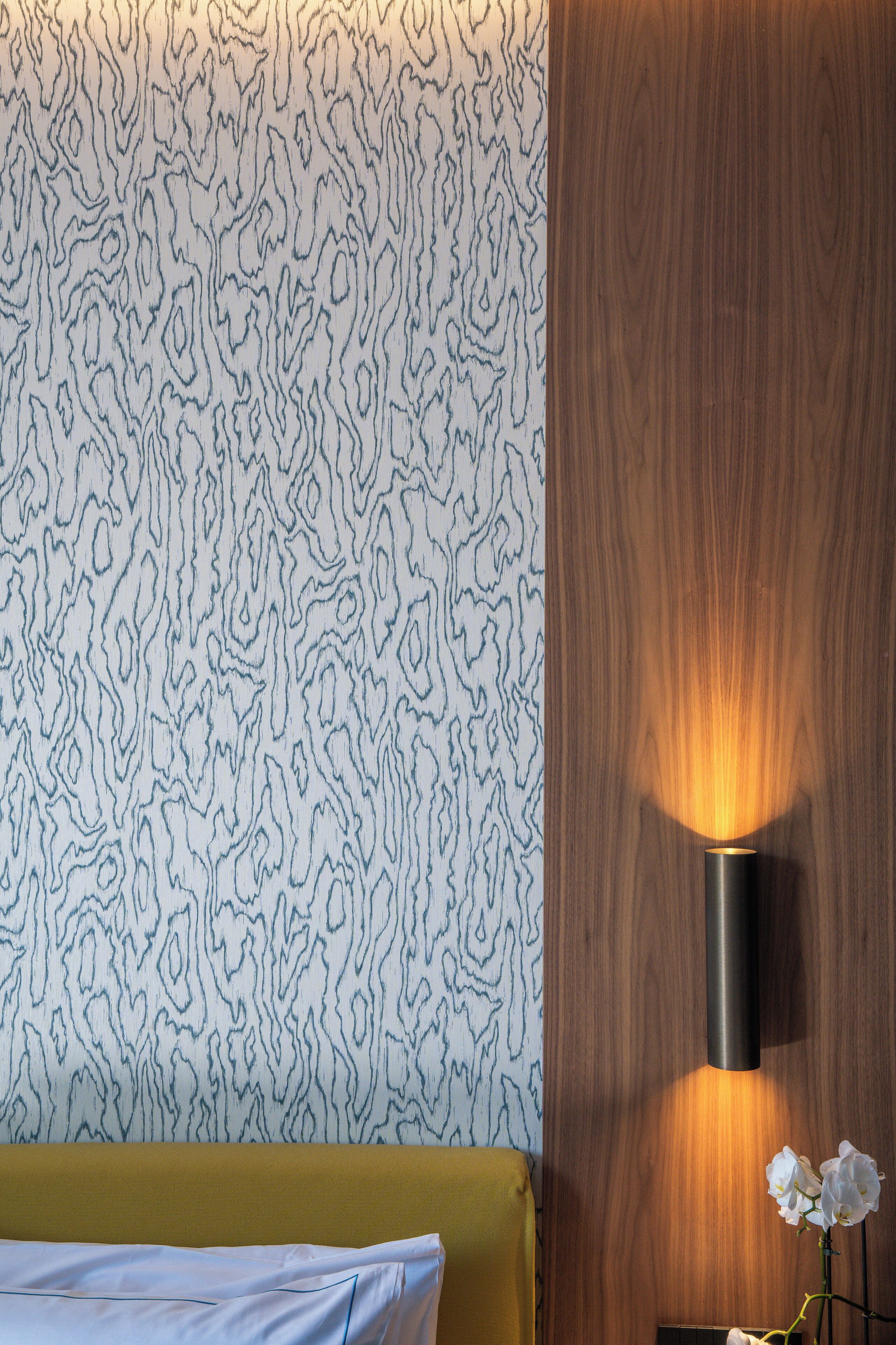Una habitación con papel tapiz estampado, un panel de madera, una lámpara de pared y una cama con cabecera amarilla.