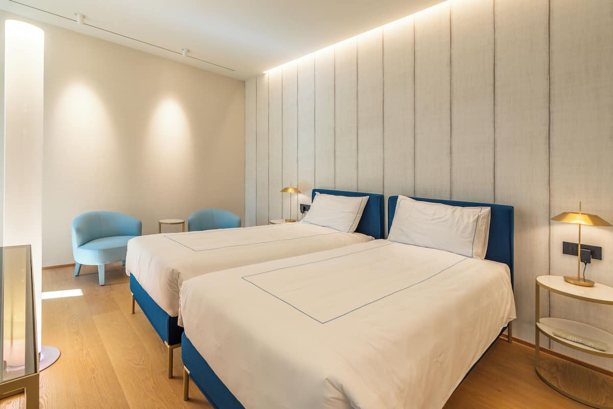 A modern hotel room with two beds, a blue armchair, a small round table, and a wall-mounted lamp.