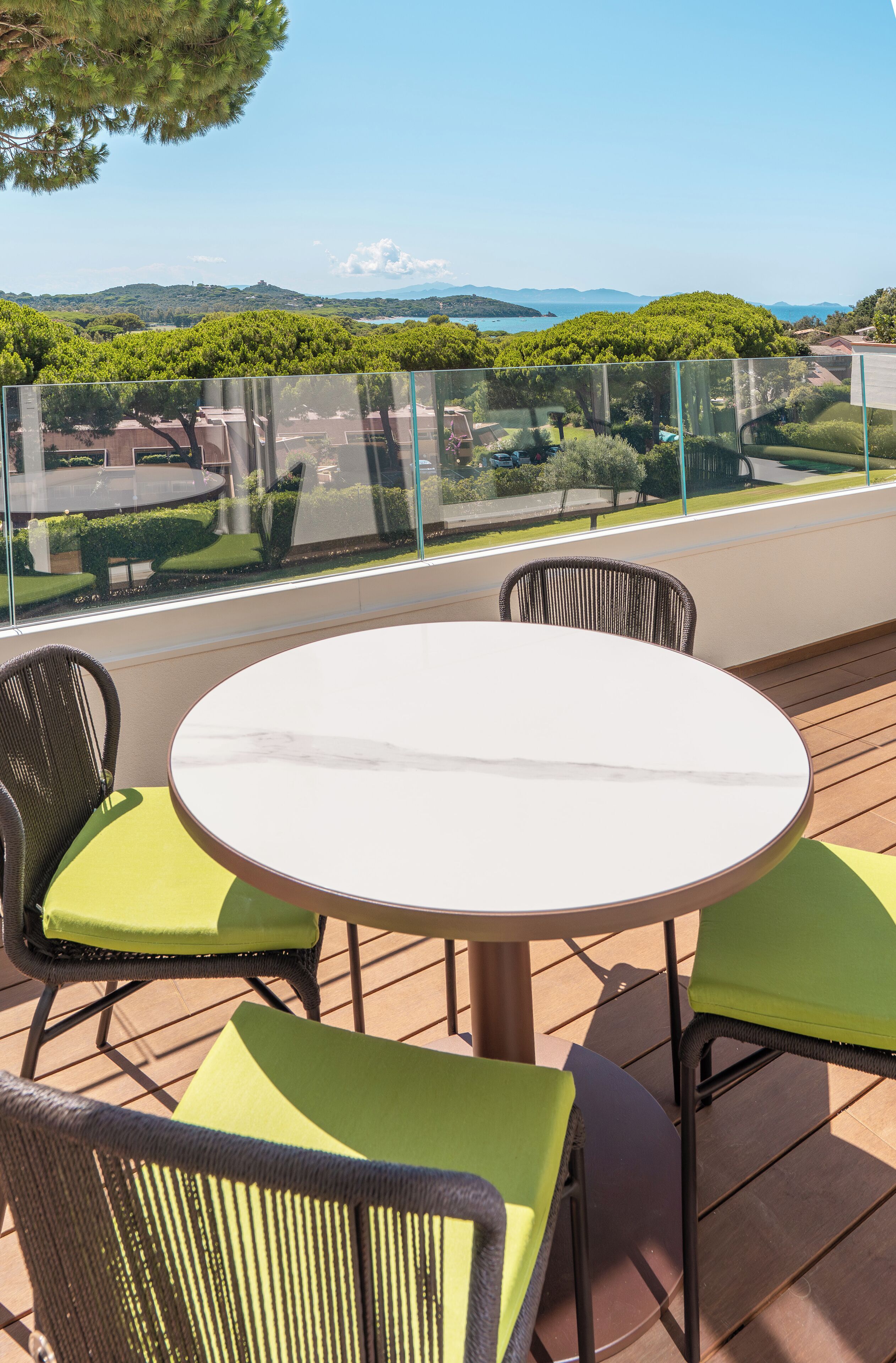 Una mesa redonda con cuatro sillas, baranda de vidrio y vista a un paisaje verde con colinas y una masa de agua.