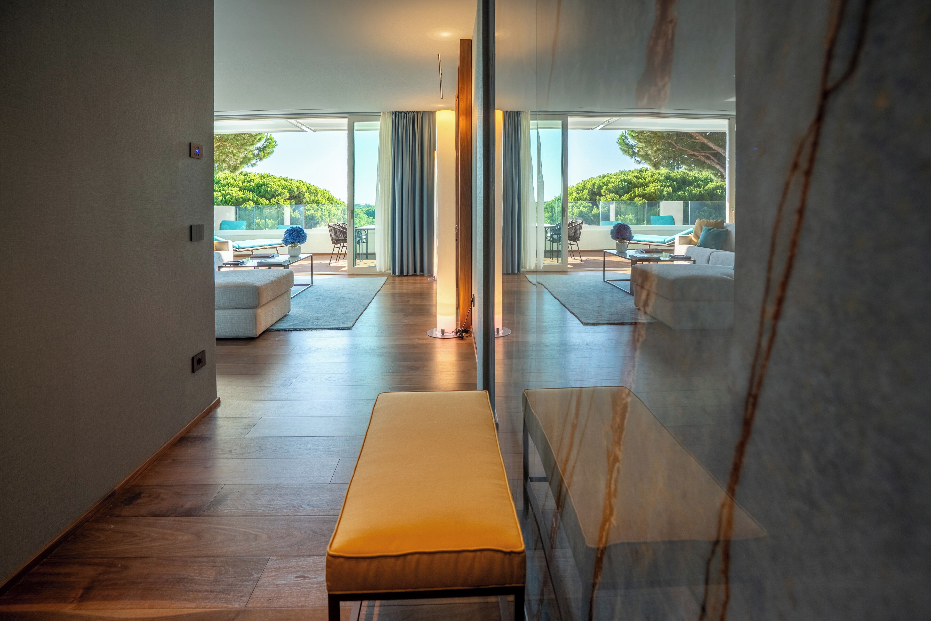 A modern interior with a bench, a glass partition, and a view of an outdoor seating area.