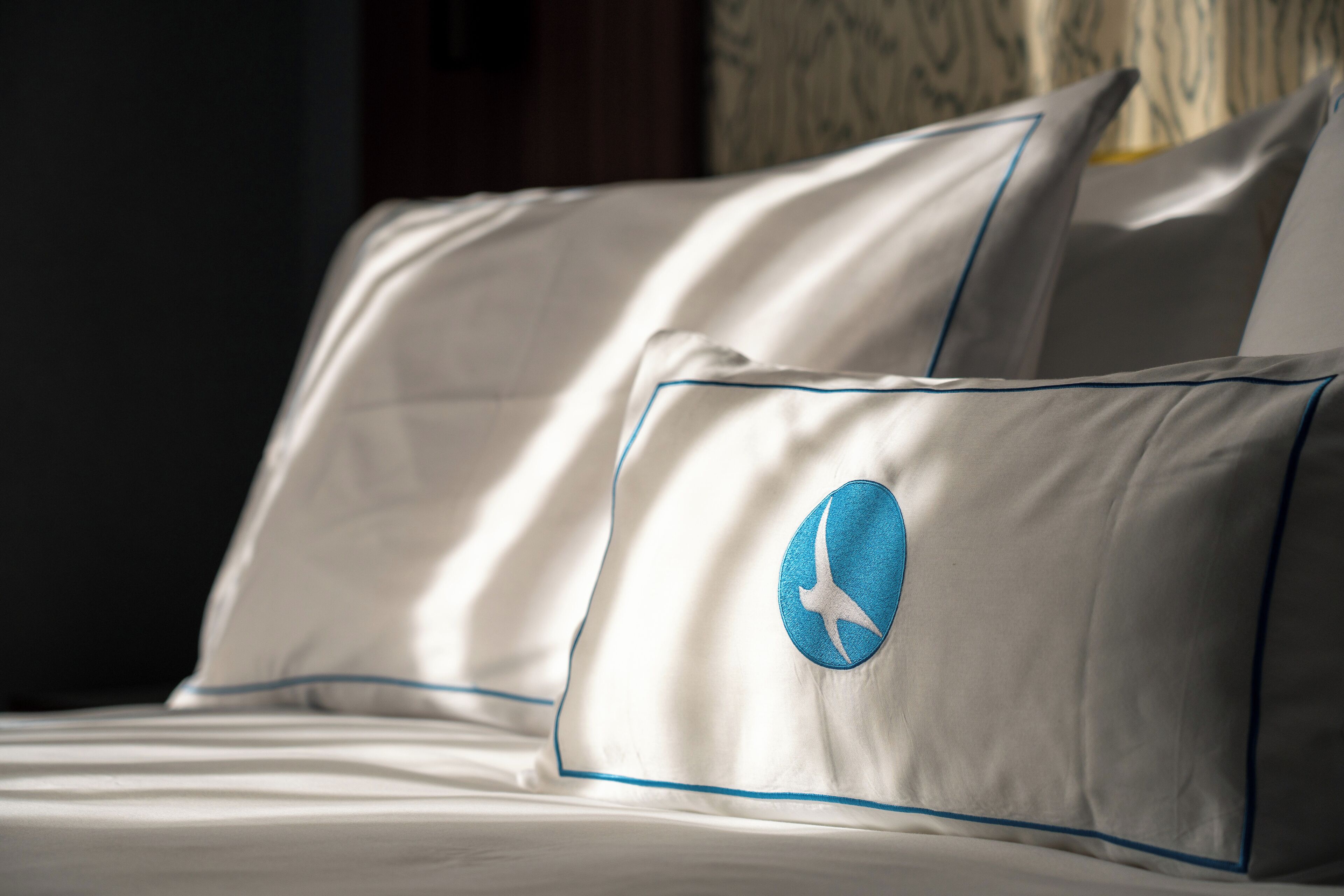 Una cama con ropa de cama y almohadas blancas, una con un logotipo bordado en azul.
