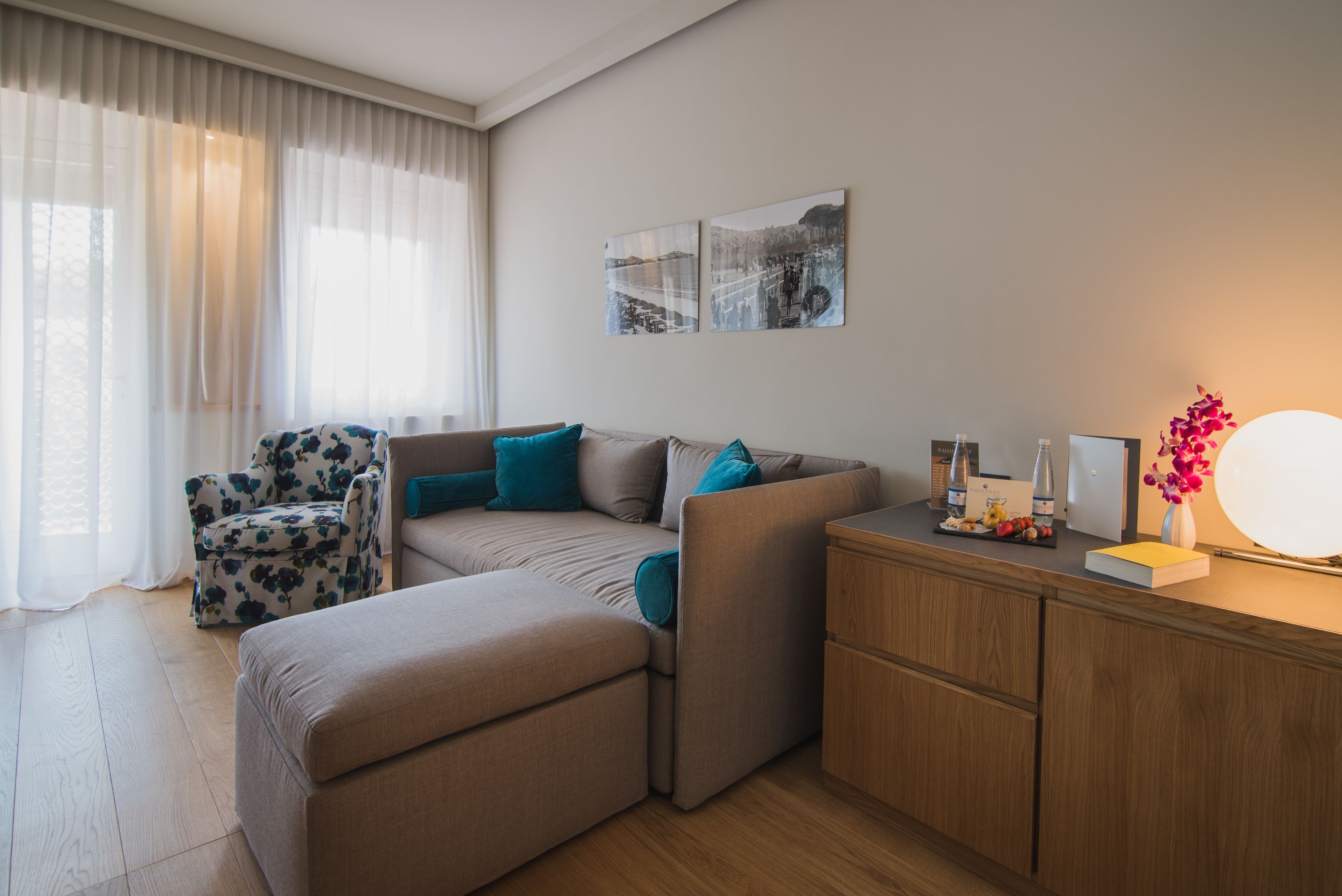 Un salon moderne avec un canapé, un pouf et un fauteuil à motifs floraux. On y trouve deux cadres accrochés au mur et une petite table d’appoint en bois, sur laquelle sont posés une lampe et des objets décoratifs.