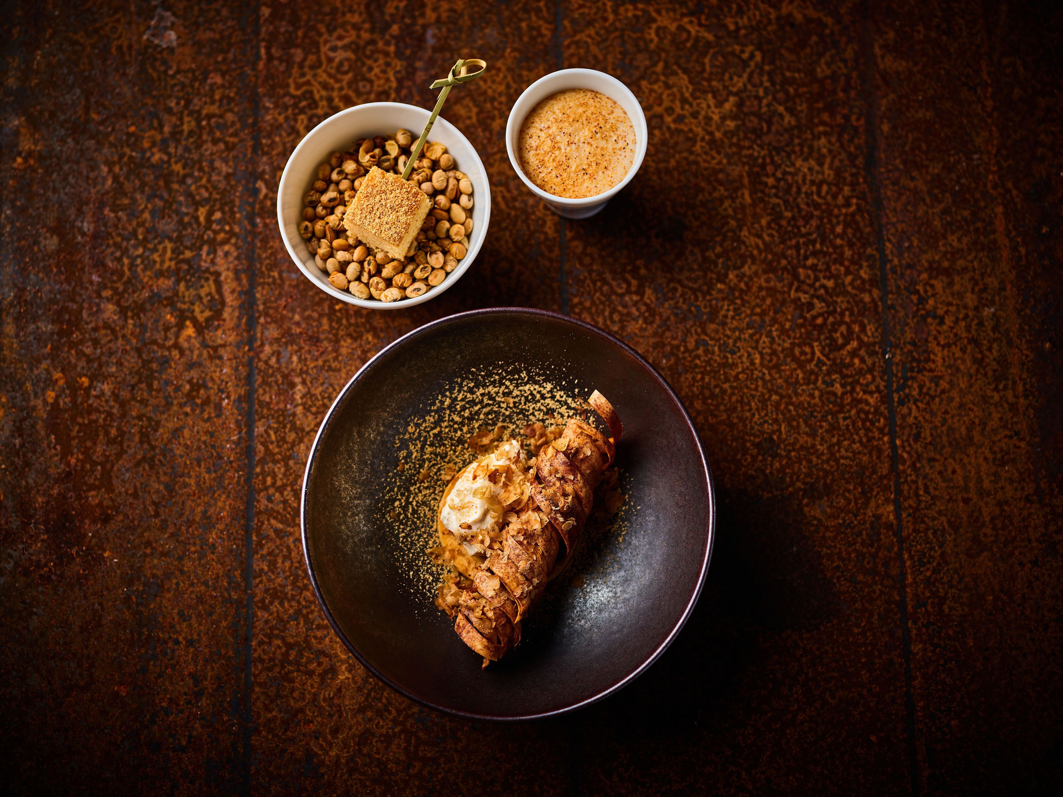A dark brown textured surface with a plate containing a food item, two bowls with nuts and a spice, and a small bowl with a liquid.
