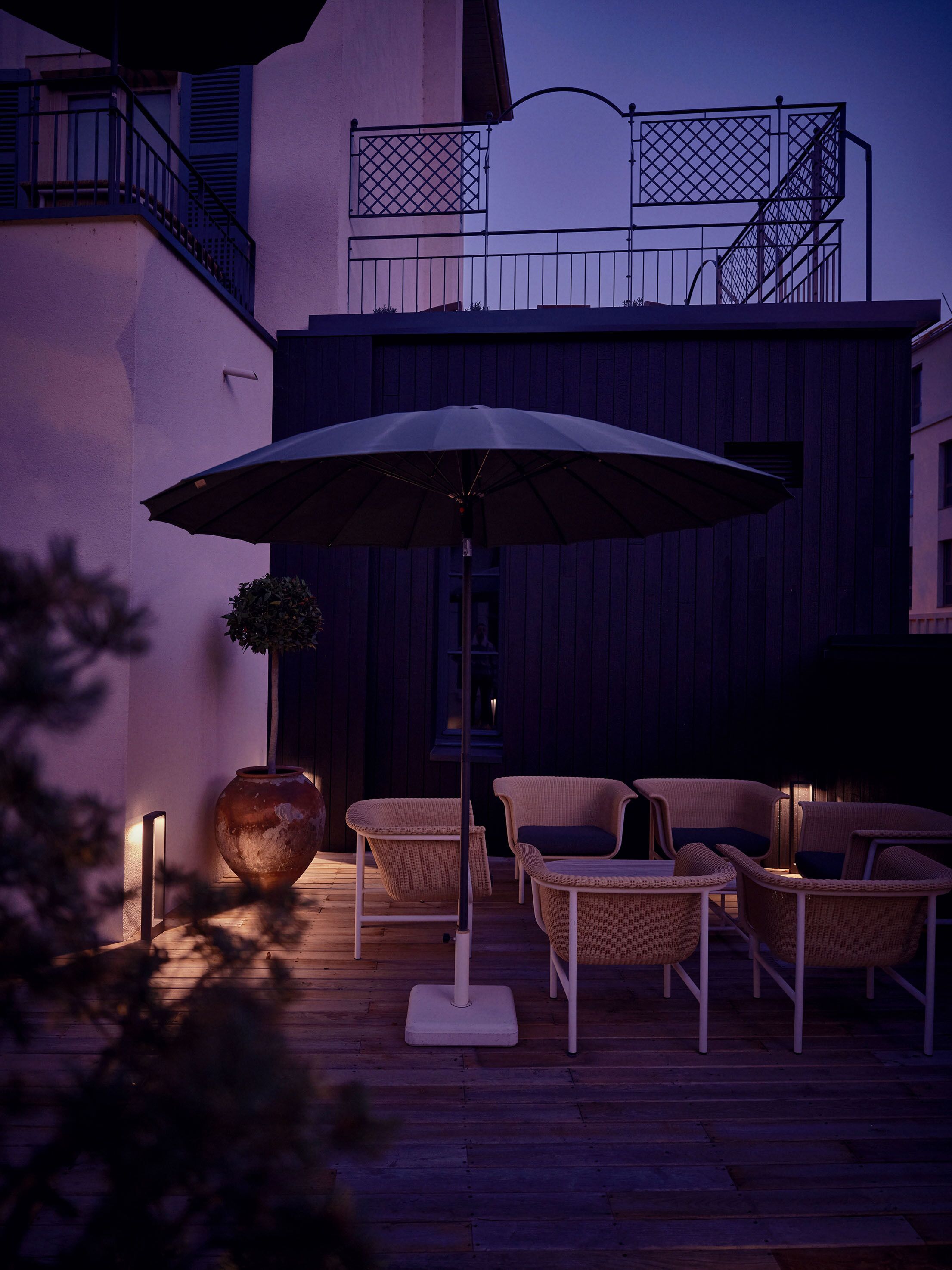 Terrassa exterior amb mobles de vimet i un parasol gran, amb un edifici i un balcó al fons.