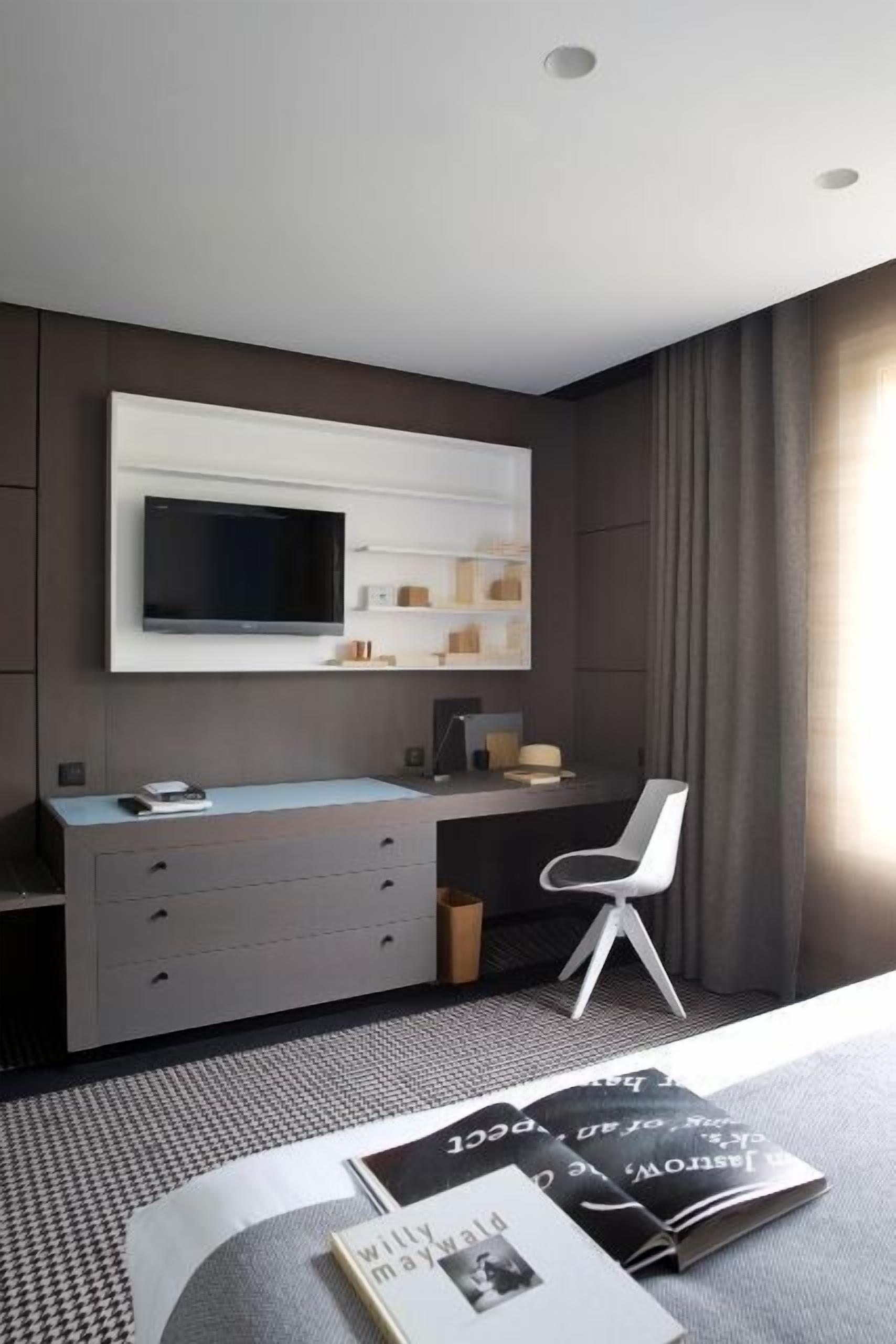 Une chambre moderne avec un grand téléviseur à écran plat fixé au mur, un bureau avec des tiroirs, une chaise et un magazine posé sur le sol.