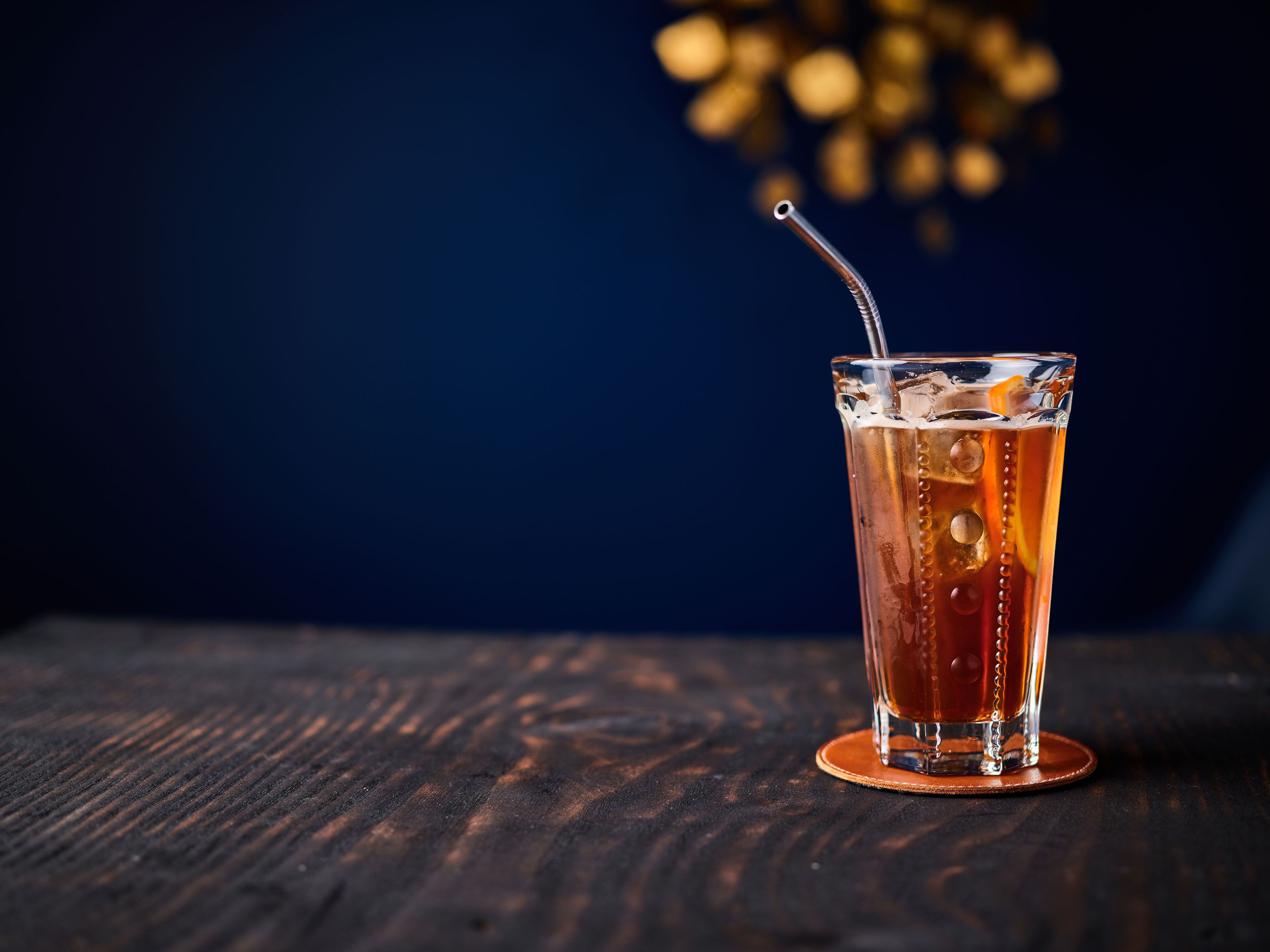 A glass of amber-colored beverage with ice cubes and a straw on a coaster, placed on a wooden surface.