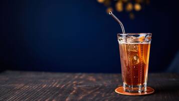 A glass of amber-colored beverage with ice cubes and a straw on a coaster, placed on a wooden surface.