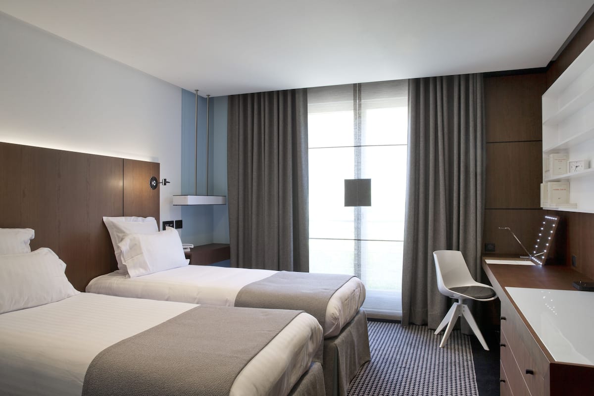 A hotel room with two beds, a desk, a chair, a vanity with a mirror, and a large window with curtains.
