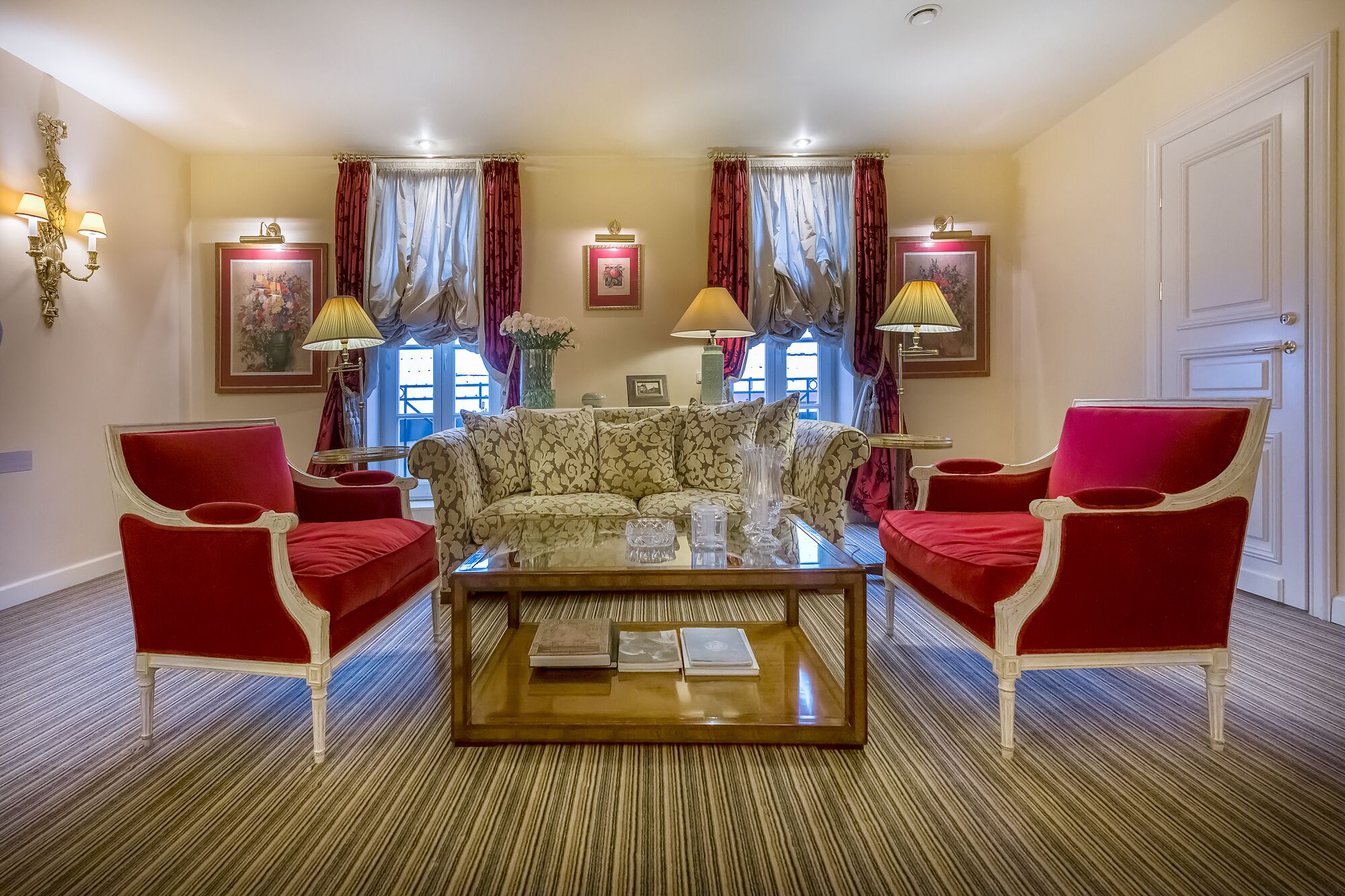 Un salon avec un canapé à motifs, des fauteuils rouges, une table basse en verre et une grande fenêtre avec des rideaux.