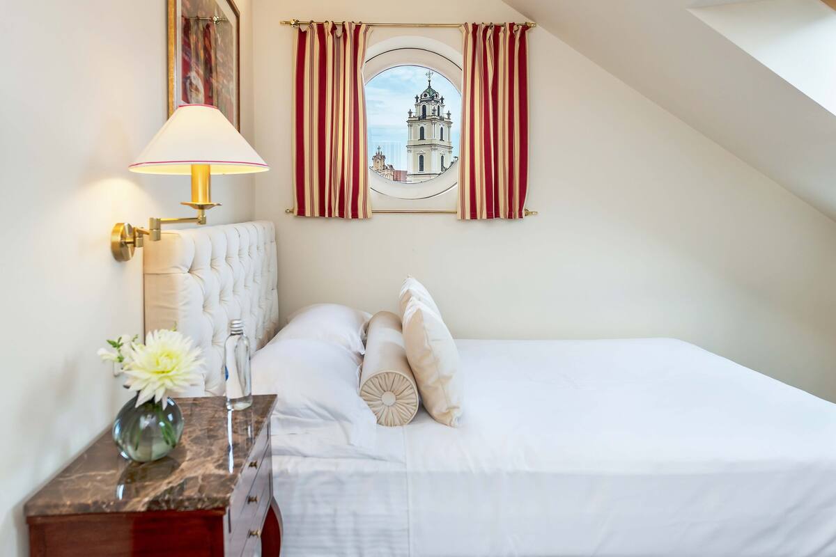 A bedroom with a bed, a nightstand, a lamp, a vase with flowers, and a window with curtains.