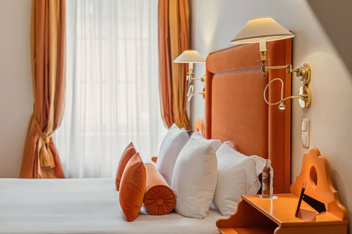 A hotel room with a bed, pillows, a nightstand, a lamp, and curtains.