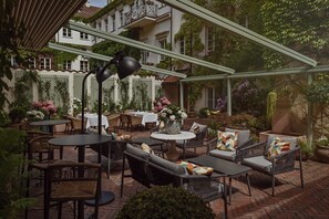 Een terras buiten met wicker meubels, ronde tafels en diverse potplanten.