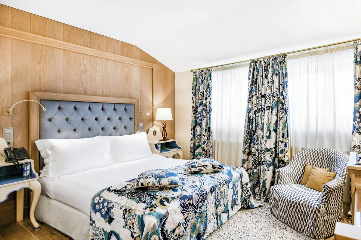 A hotel room with a tufted headboard bed, a patterned bedspread, a patterned armchair, and a nightstand with a lamp and phone.