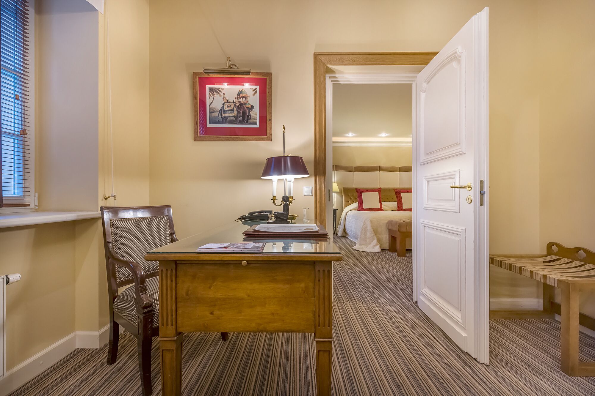 Ein Hotelzimmer mit einem Holztisch, einer Lampe, einem gerahmten Bild an der Wand und einem Bett mit einem roten Kissen.