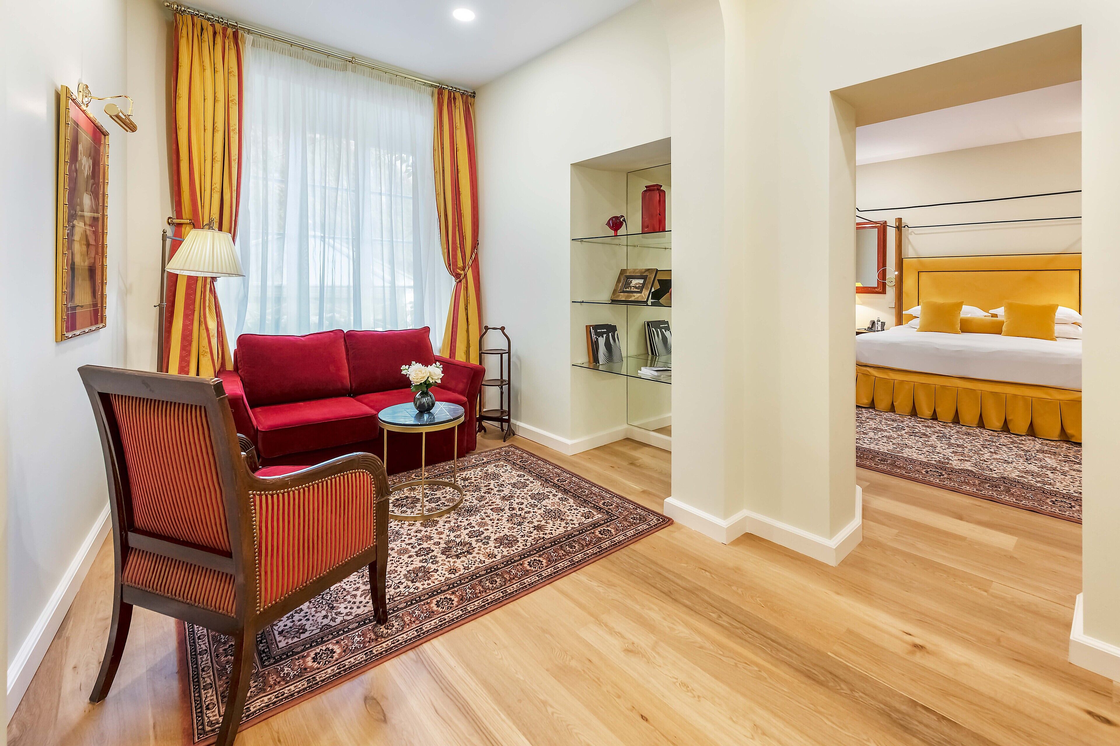 Une chambre d’hôtel avec un canapé rouge, une petite table ronde avec une lampe, une chaise en bois, une vitrine et un lit avec une tête de lit jaune.