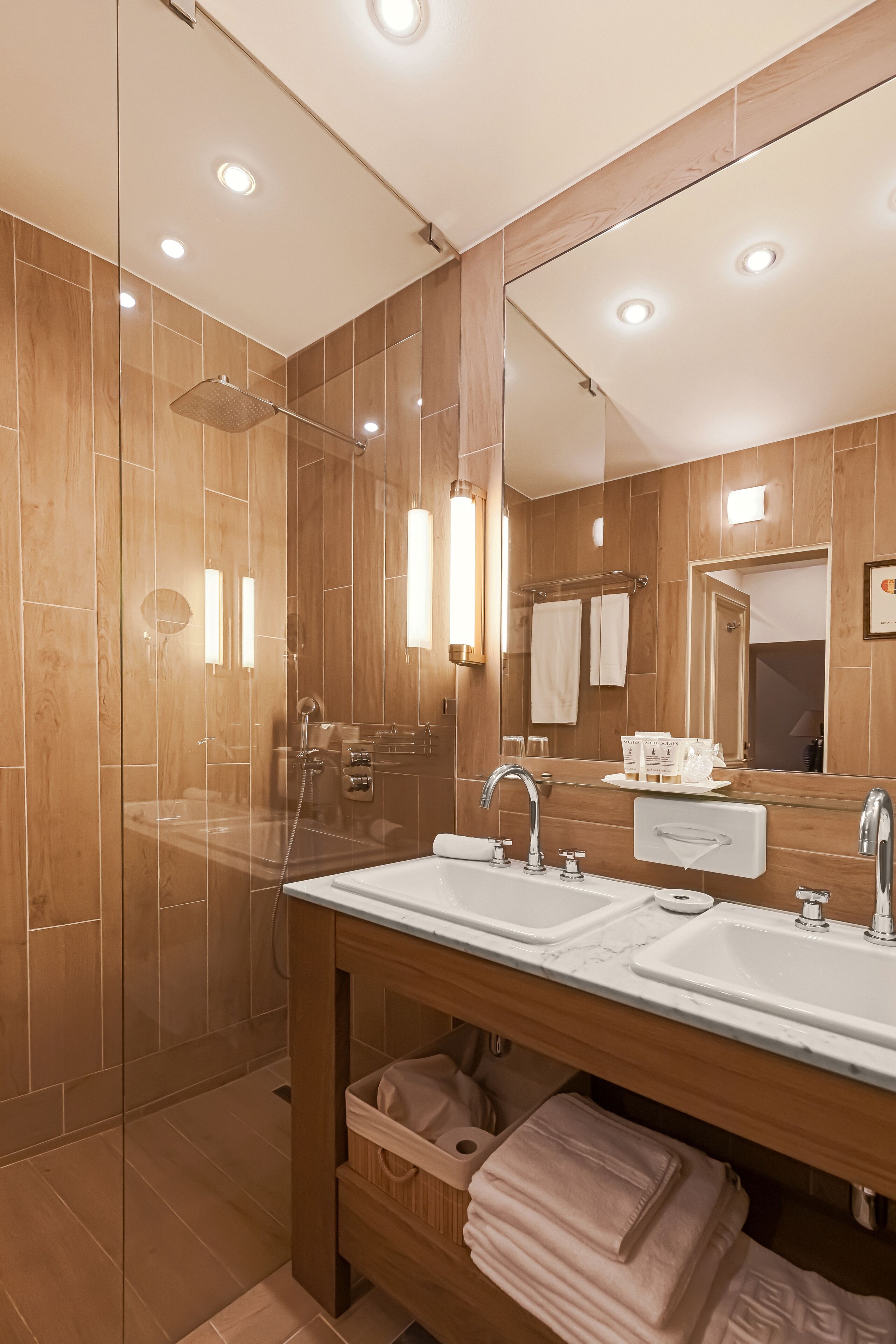 Un baño moderno con ducha envuelta en vidrio, tocador de madera con dos lavamanos y un espejo grande.