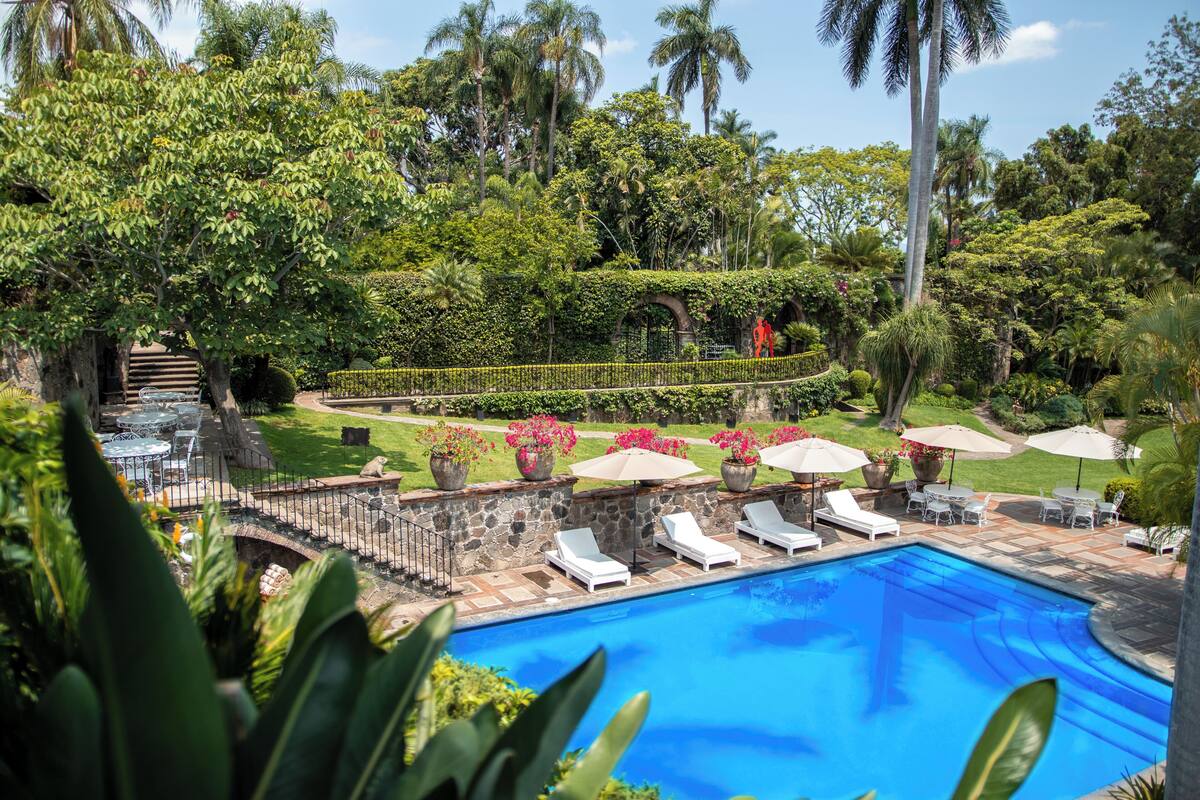 Una piscina con sillones de descanso, rodeada de abundante vegetación y un puente de piedra.