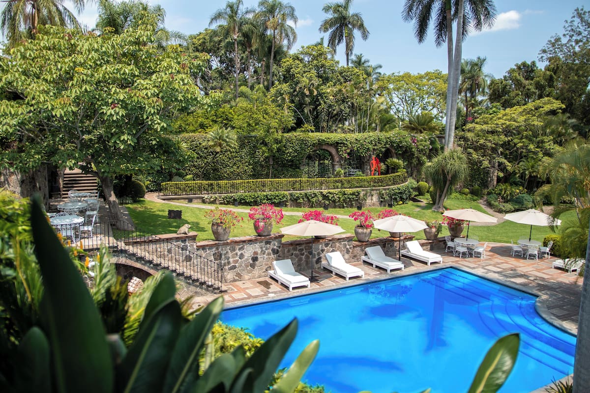 A swimming pool with lounge chairs, surrounded by lush greenery and a stone bridge.