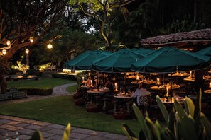 Een terras met tafels en groene parasols, omgeven door bomen en plantenpotten.