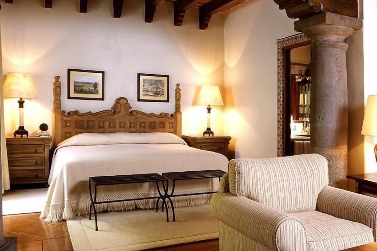 Un dormitorio con una cama de madera, dos mesitas de noche con lámparas, un banco, un sillón a rayas y una columna de piedra.