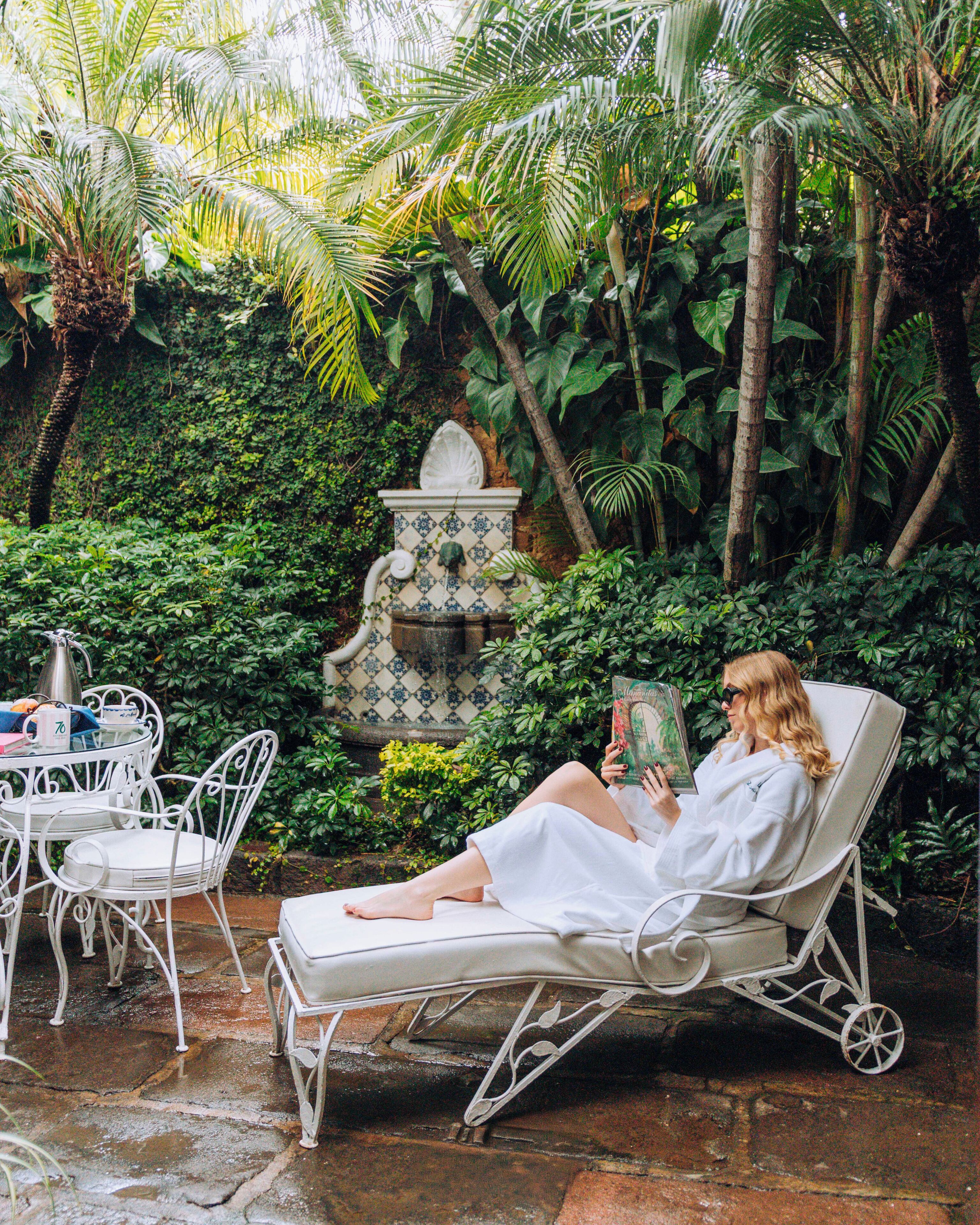 Una mujer leyendo un libro en una silla lounge blanca, rodeada de exuberante follaje y una fuente decorativa.
