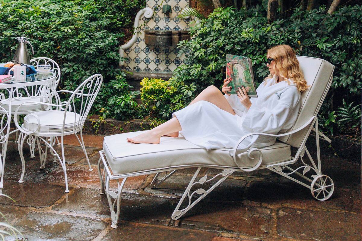 Una mujer leyendo un libro en una silla lounge blanca, rodeada de exuberante follaje y una fuente decorativa.
