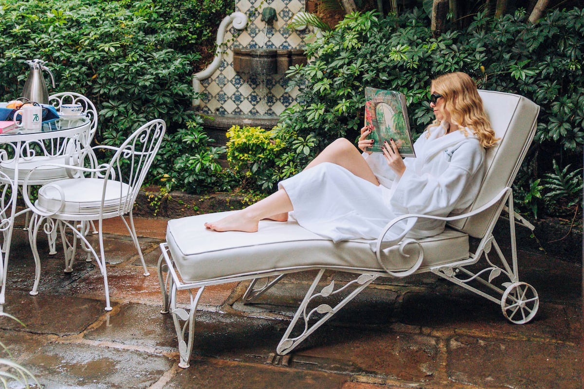 A woman reading a book in a white lounge chair, surrounded by lush greenery and a decorative fountain.