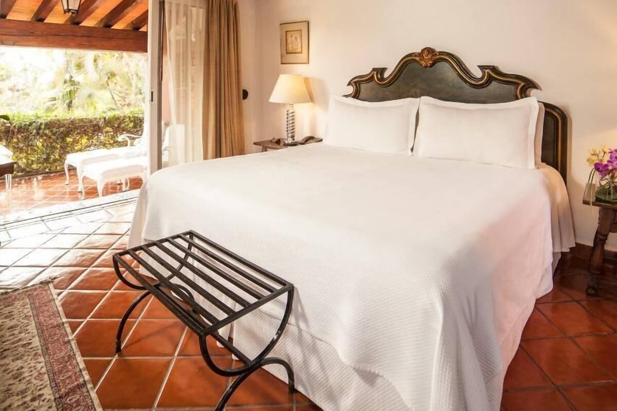 Un dormitorio con cama, mesita de noche, lámpara, silla y vistas al exterior.