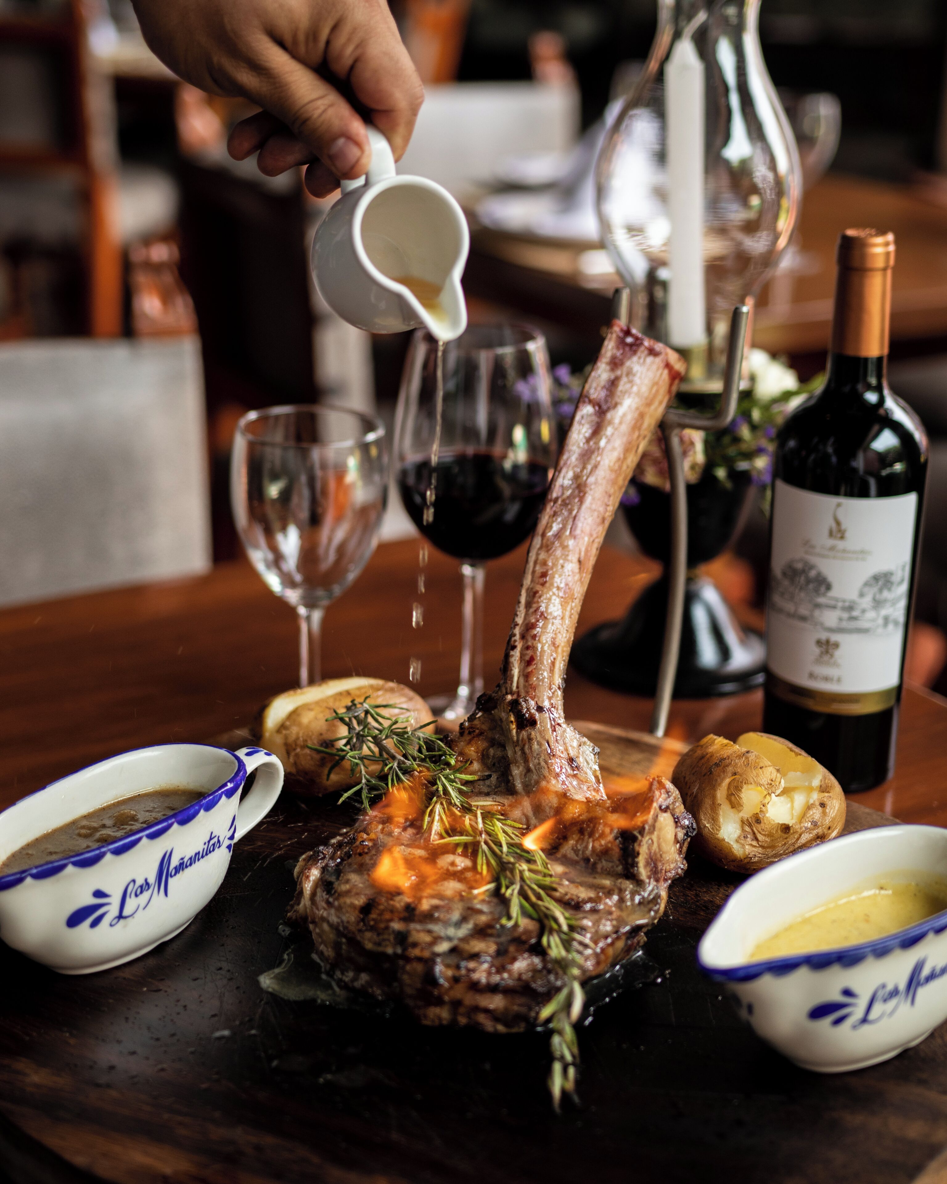Un plato de filete con romero, acompañado de una botella de vino, una copa de vino tinto y dos salseras con salsa.