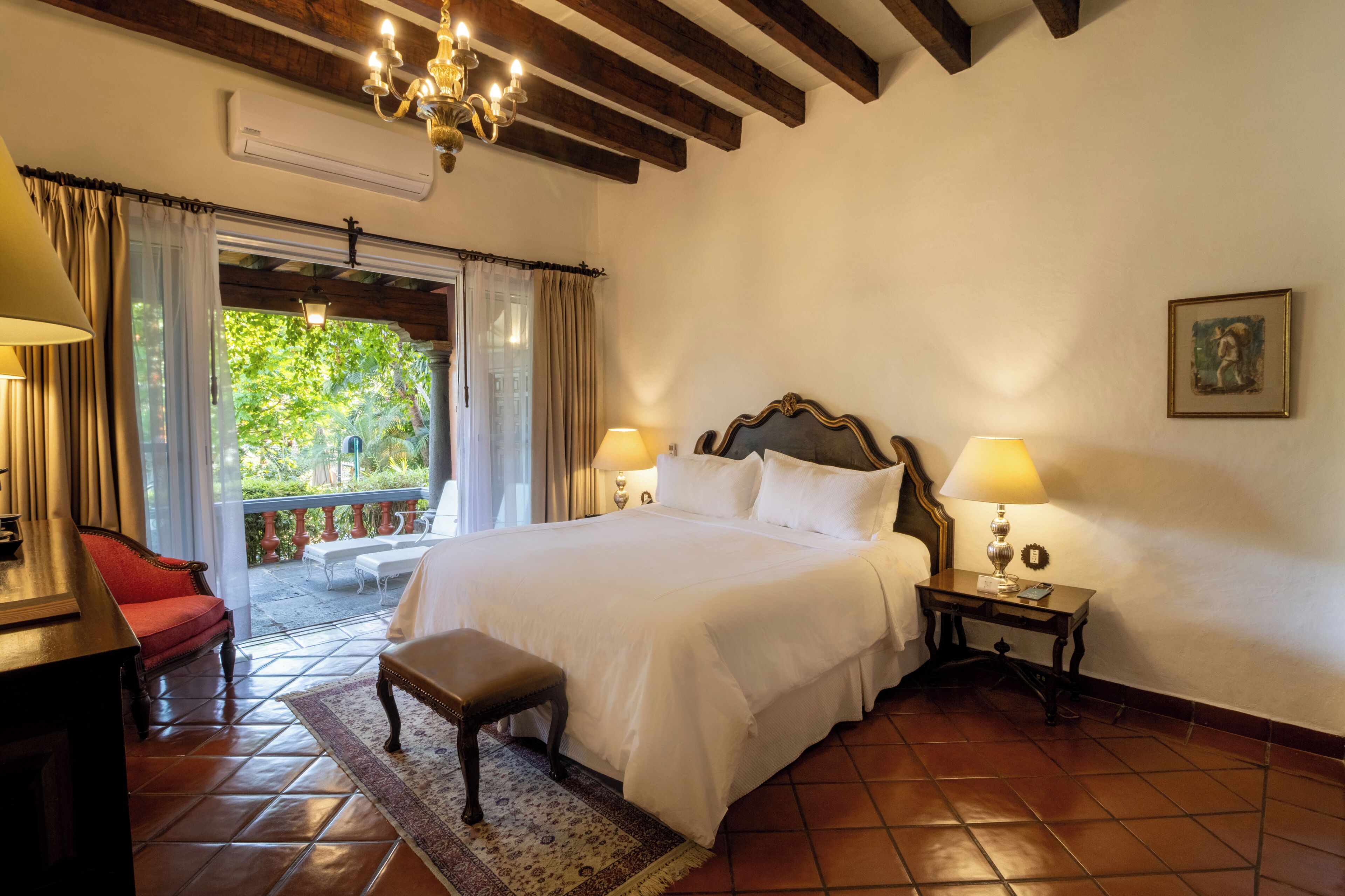 A hotel room with a large bed, two bedside lamps, a small table, a bench, and a view of greenery through a window.