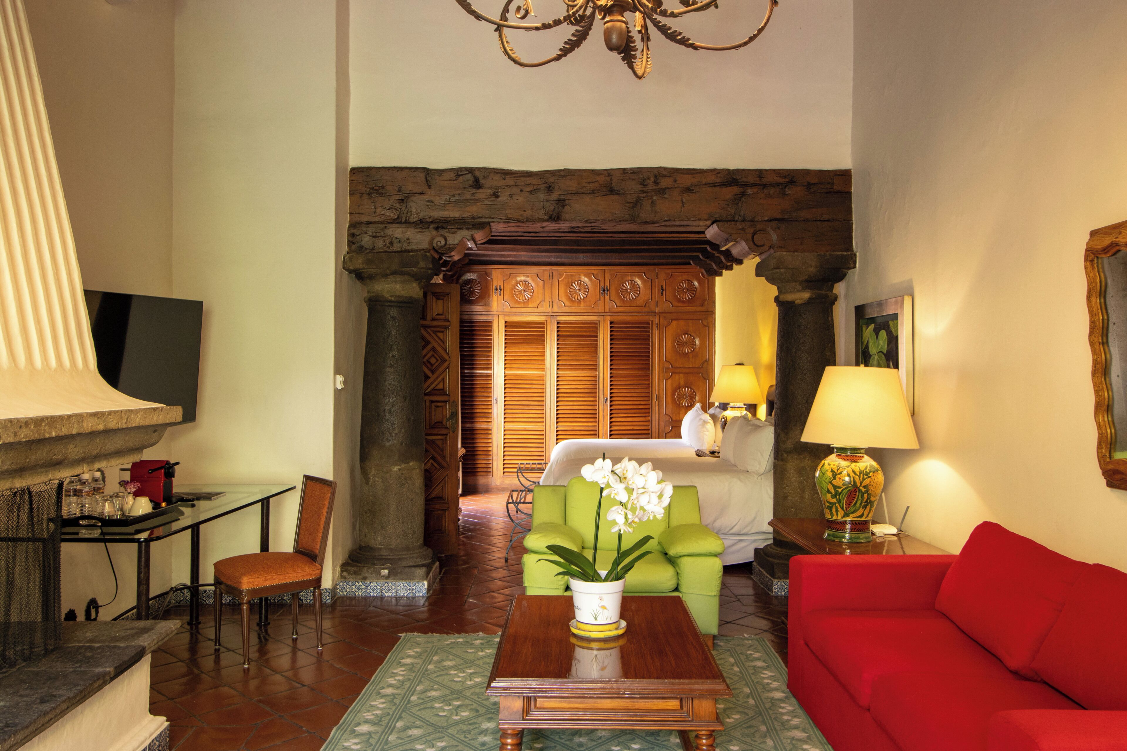 Un vestíbulo de hotel con un mostrador de recepción de madera, un sillón verde, un sofá rojo, una mesa de centro de madera y una araña.