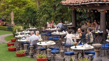 Un área de comedor al aire libre con varias mesas y sillas, rodeada de exuberante vegetación y una estructura de techo de tejas.