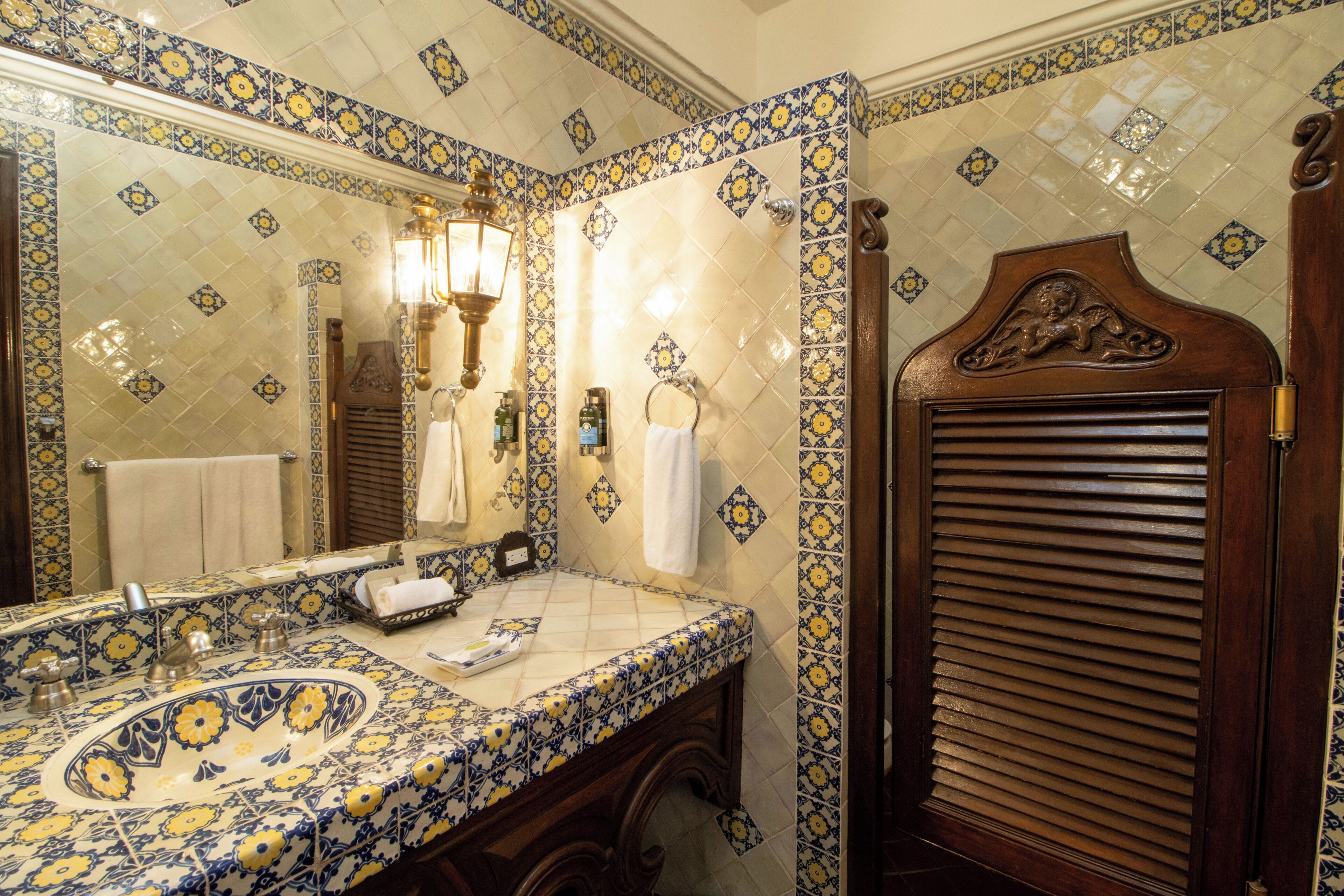 Un baño con salpicadero de azulejos estampados, un lavabo con pileta decorativa, un espejo y una puerta de madera con pomo decorativo.