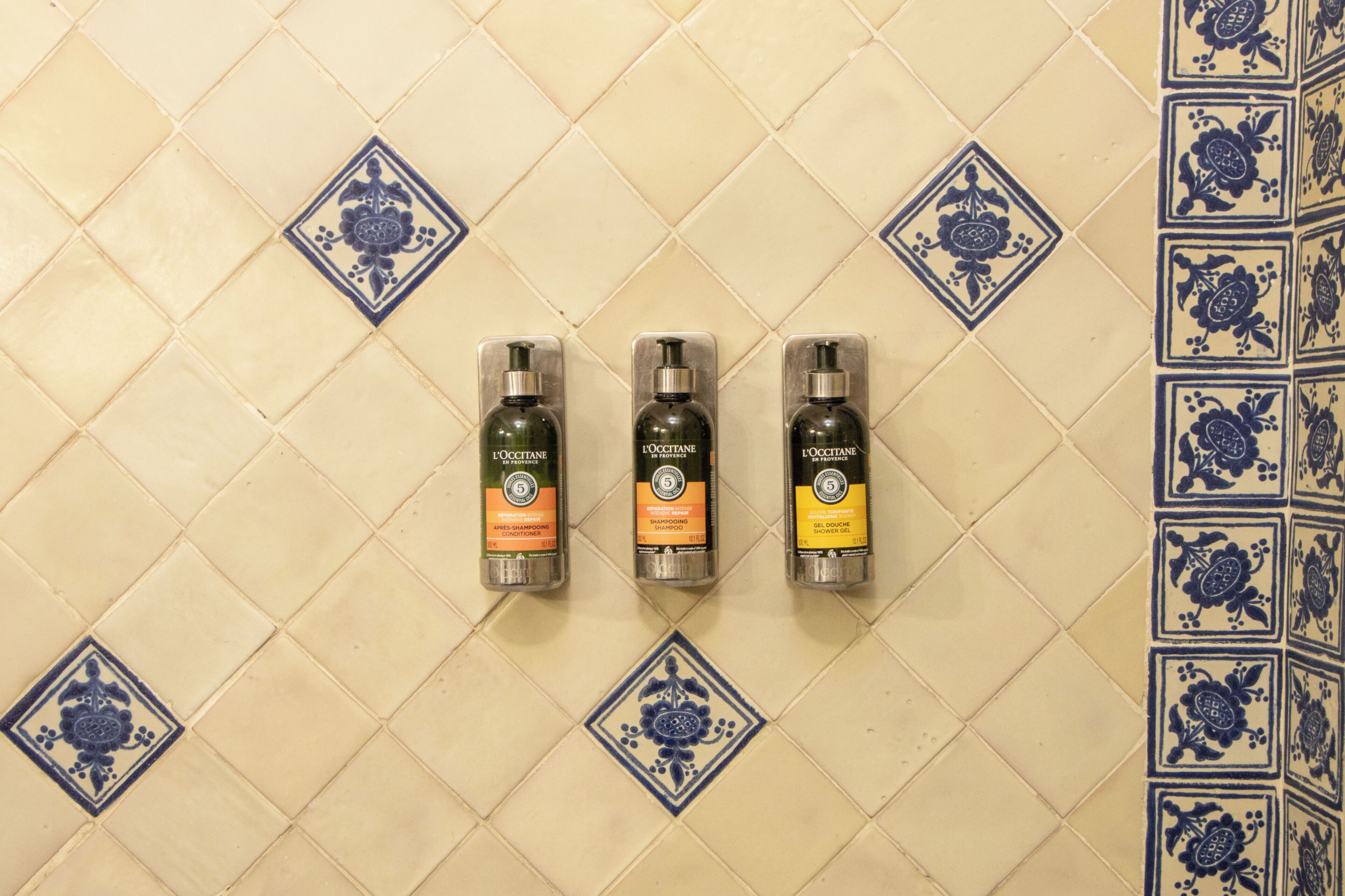 Tres botellas de champú de L'Occitane sobre una pared de azulejos con un estampado floral azul.
