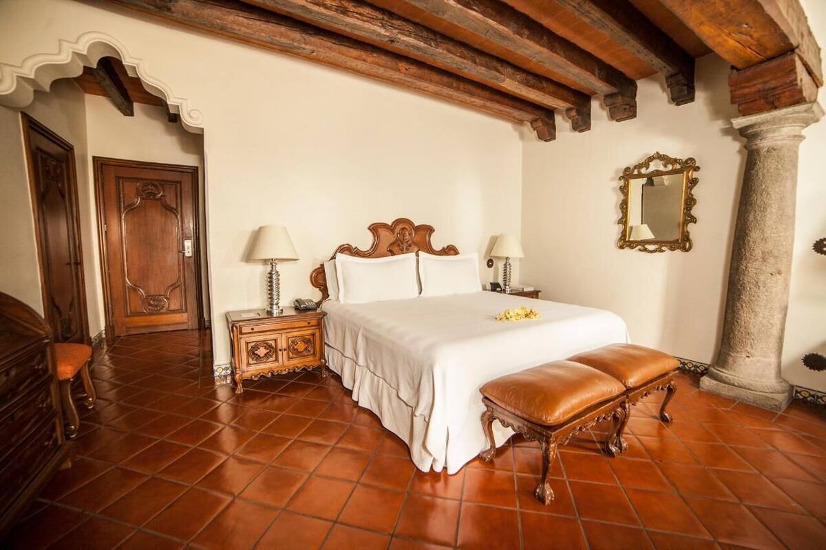Un dormitorio con una cama, mesitas de noche, un banco, un espejo y una puerta.