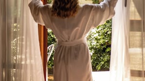 Eine Person in einem weißen Bademantel steht an einem Fenster mit transparenten Vorhängen und blickt auf einen üppigen, grünen Garten.
