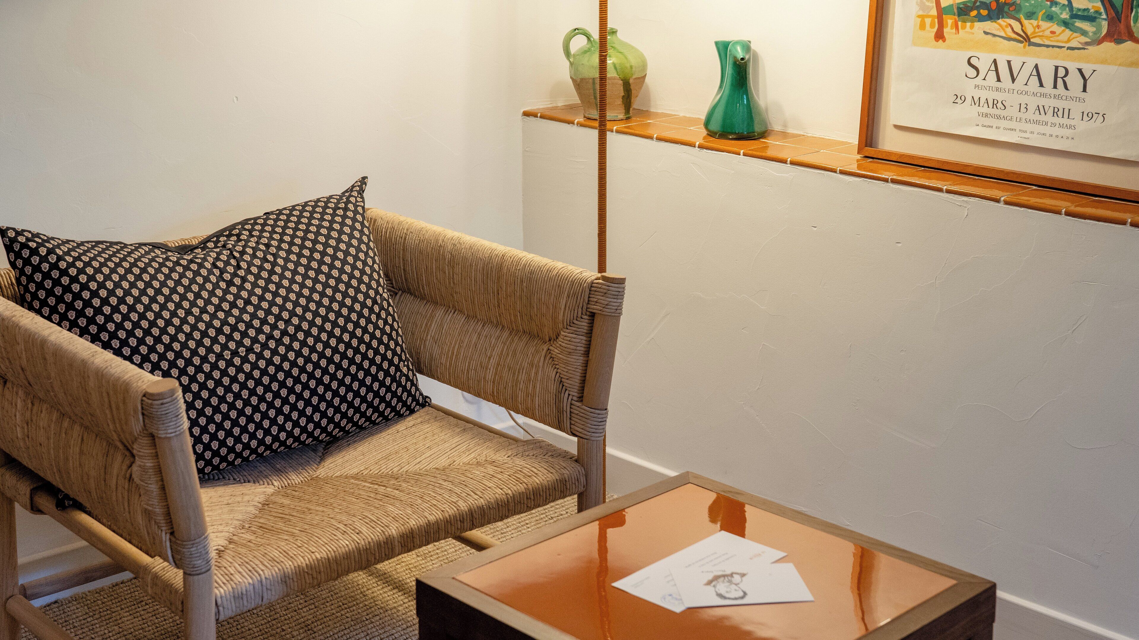 A room with a wicker sofa, a patterned pillow, a small table with papers, and a framed artwork on the wall.