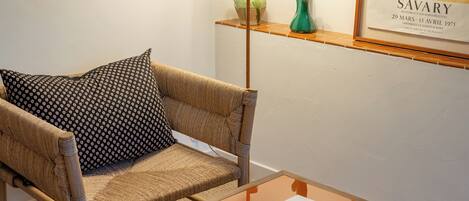 A room with a wicker sofa, a patterned pillow, a small table with papers, and a framed artwork on the wall.