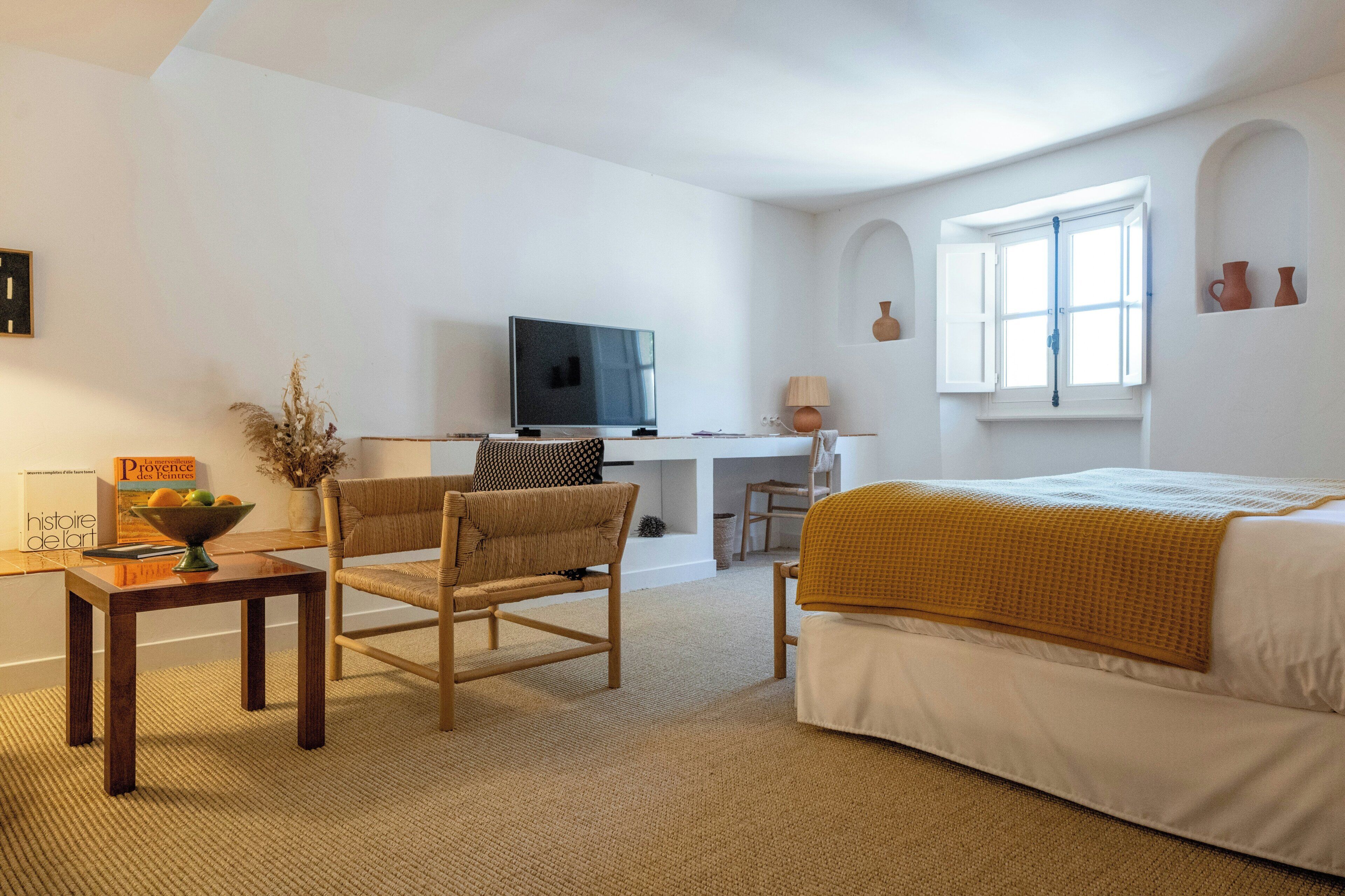A room with a bed, a desk, a television, a chair, and a small table with a bowl of fruit.