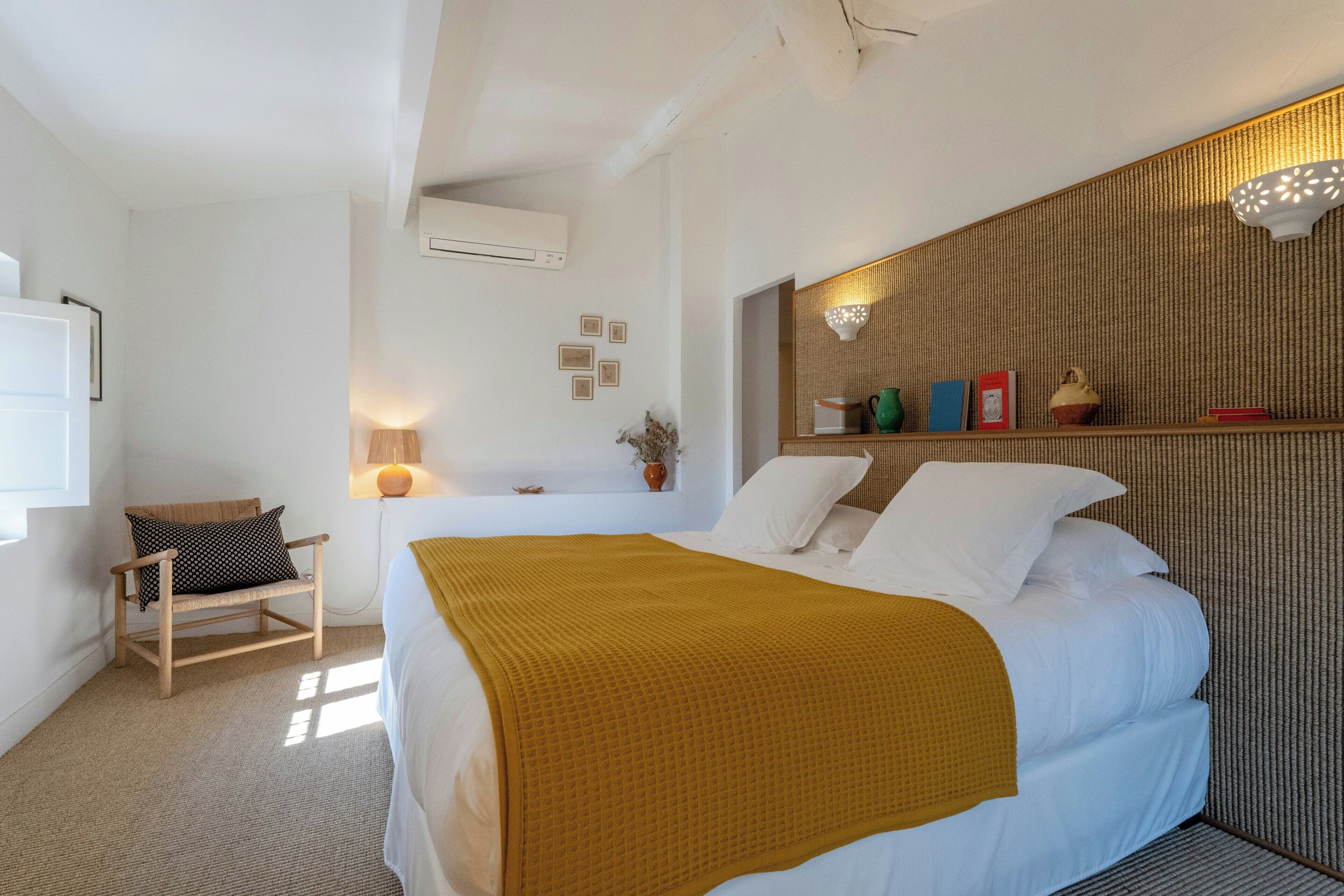 A bedroom with a bed, a chair, a bedside table with a lamp, and a wall-mounted shelf with decorative items.