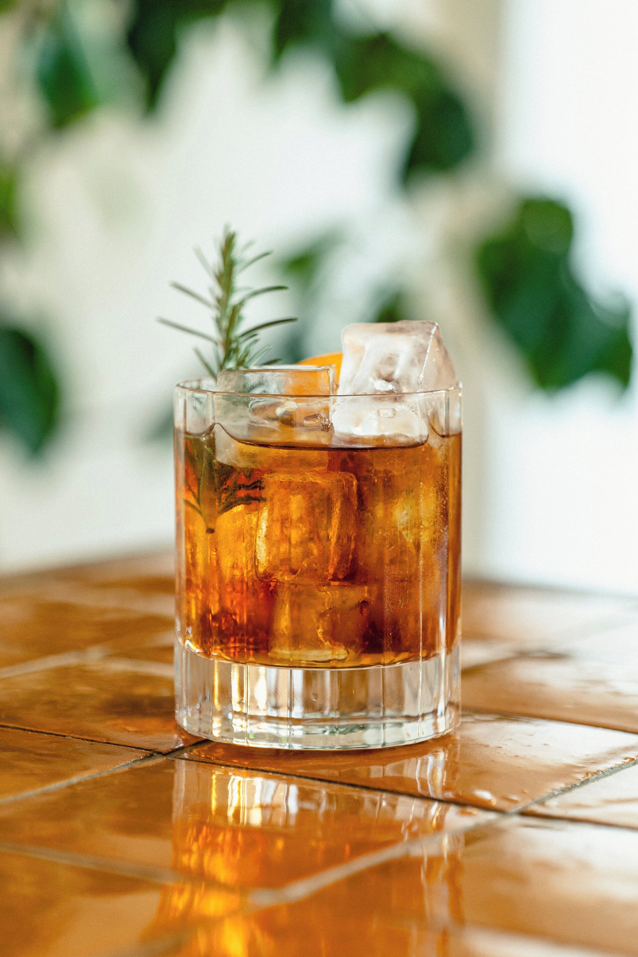 A clear glass with a beverage, ice cubes, and a sprig of rosemary on a reflective surface.