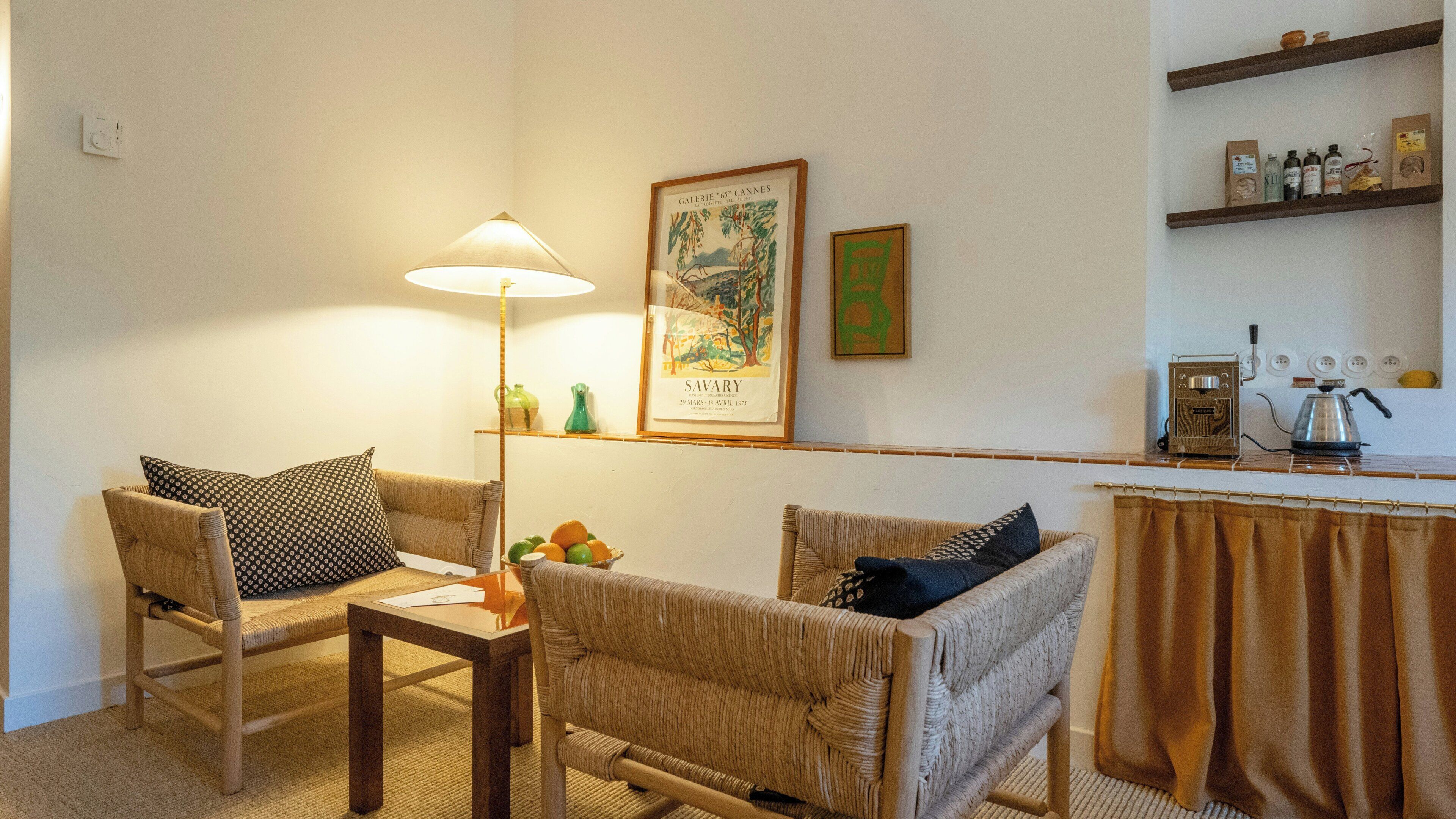 Een kamer met rieten meubels, een tafel met een lamp, een lijst met een schilderij en een keukenhoek met een koffieapparaat.