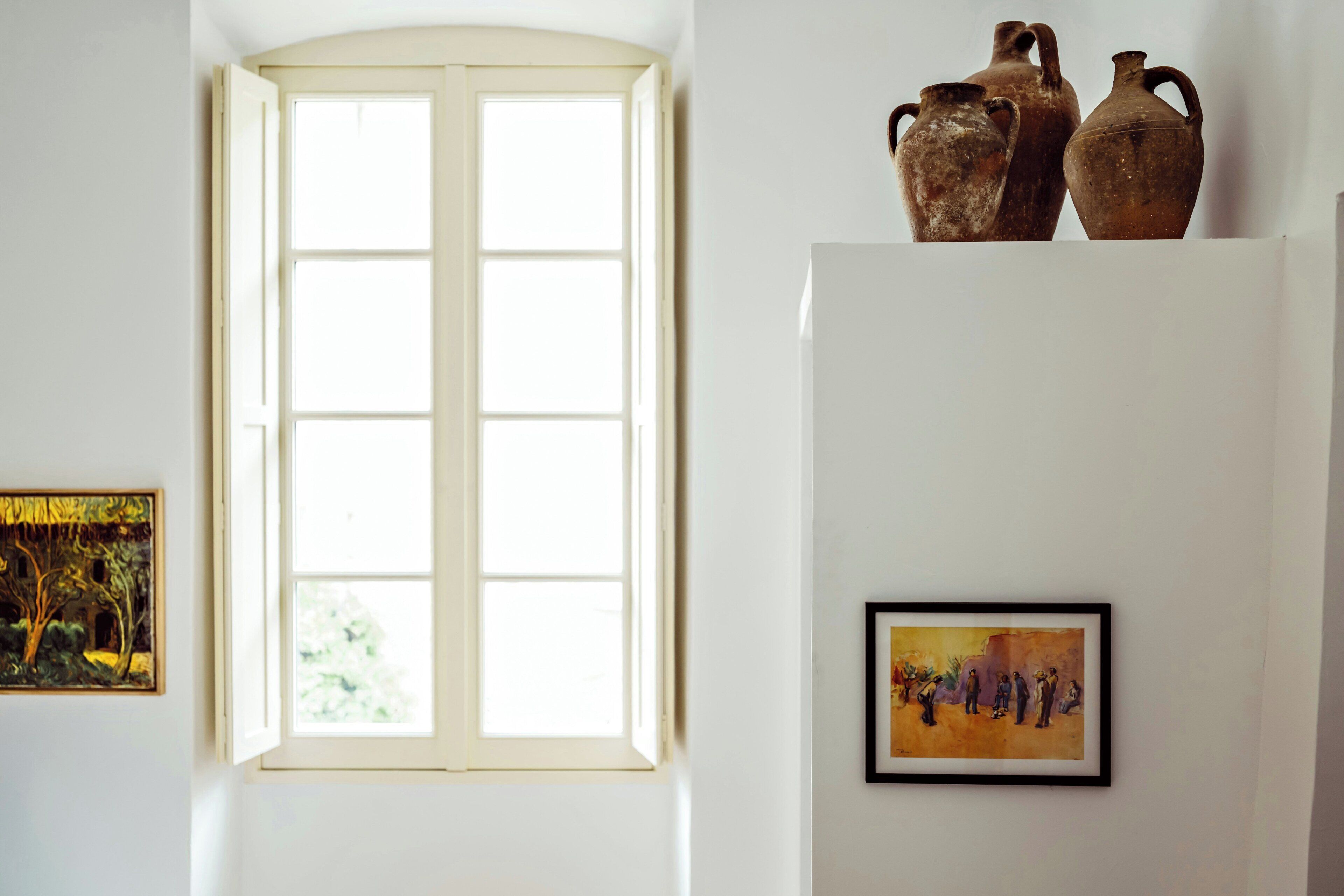 A room with a large window, a painting on the wall, and a shelf with pottery.