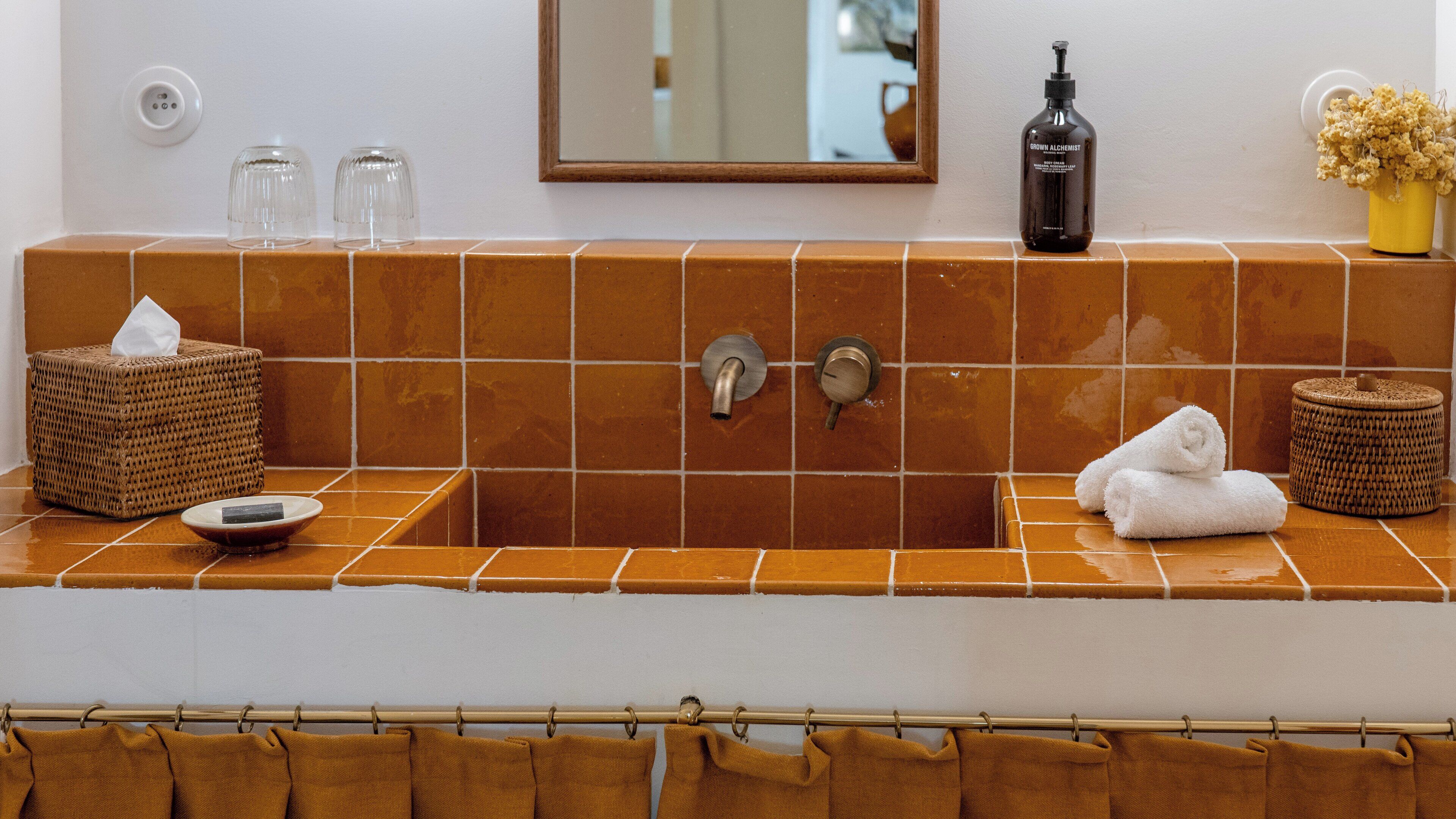 Ein Badezimmer mit einem braunen Fliesenwaschbecken, einem Spiegel, einer Shampooflasche, einer Seifenschachtel und einem Handtuch.