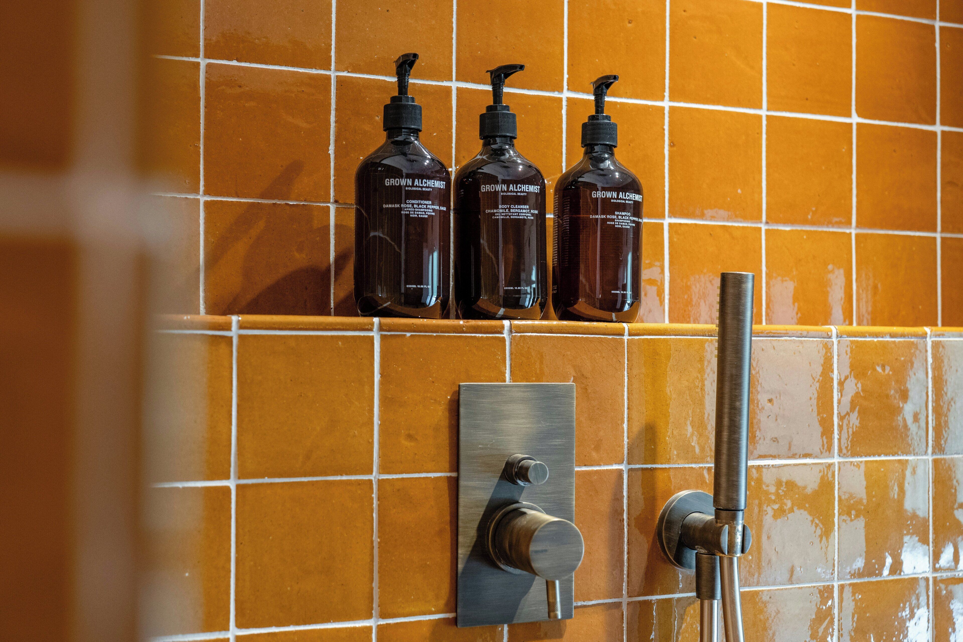 A bathroom with a tiled wall, a hand dryer, and three bottles of 'Grown Alchemist' body wash.