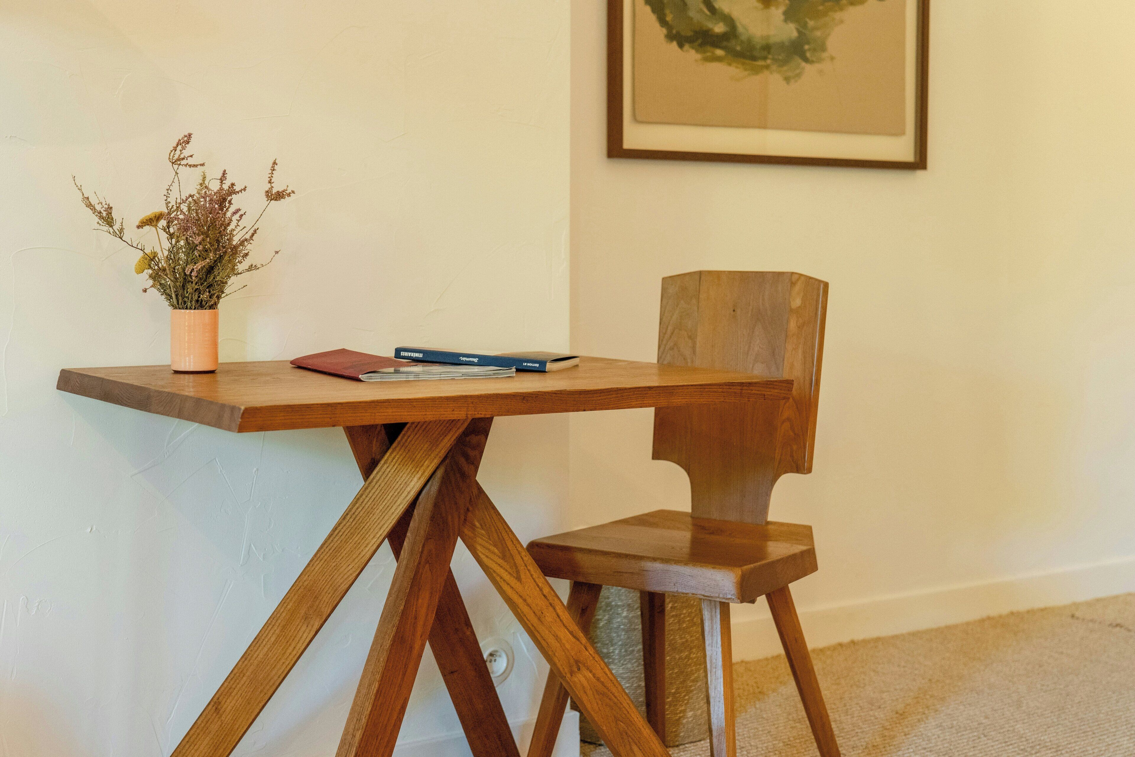A wooden table with a potted plant and a book, a wooden chair, and a framed picture on the wall.
