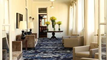 En hotellobby med et mønstret tæppe, flere sofaer, et rundt bord med blomsterarrangementer og en lysekrone.
