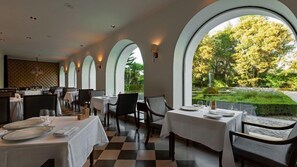 A restaurant with a checkered floor, tables set with white tablecloths, and large arched windows overlooking a garden.